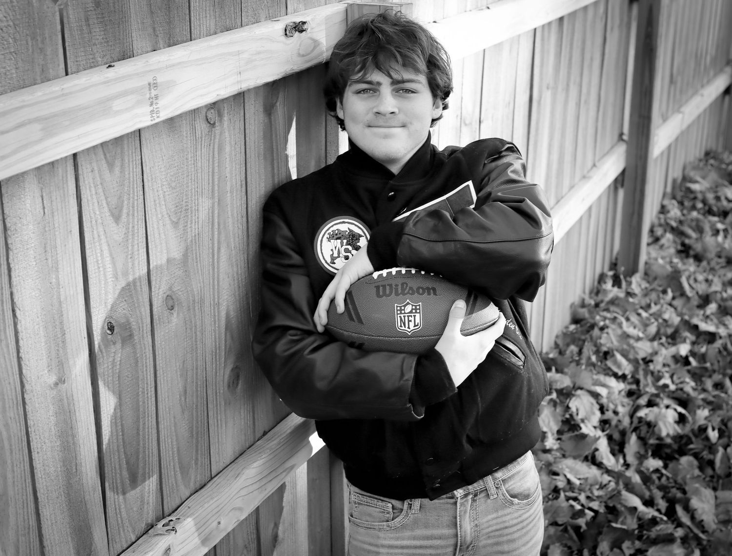 A black and white photo shows someone wearing a dark jacket leaning against a wooden fence outdoors.