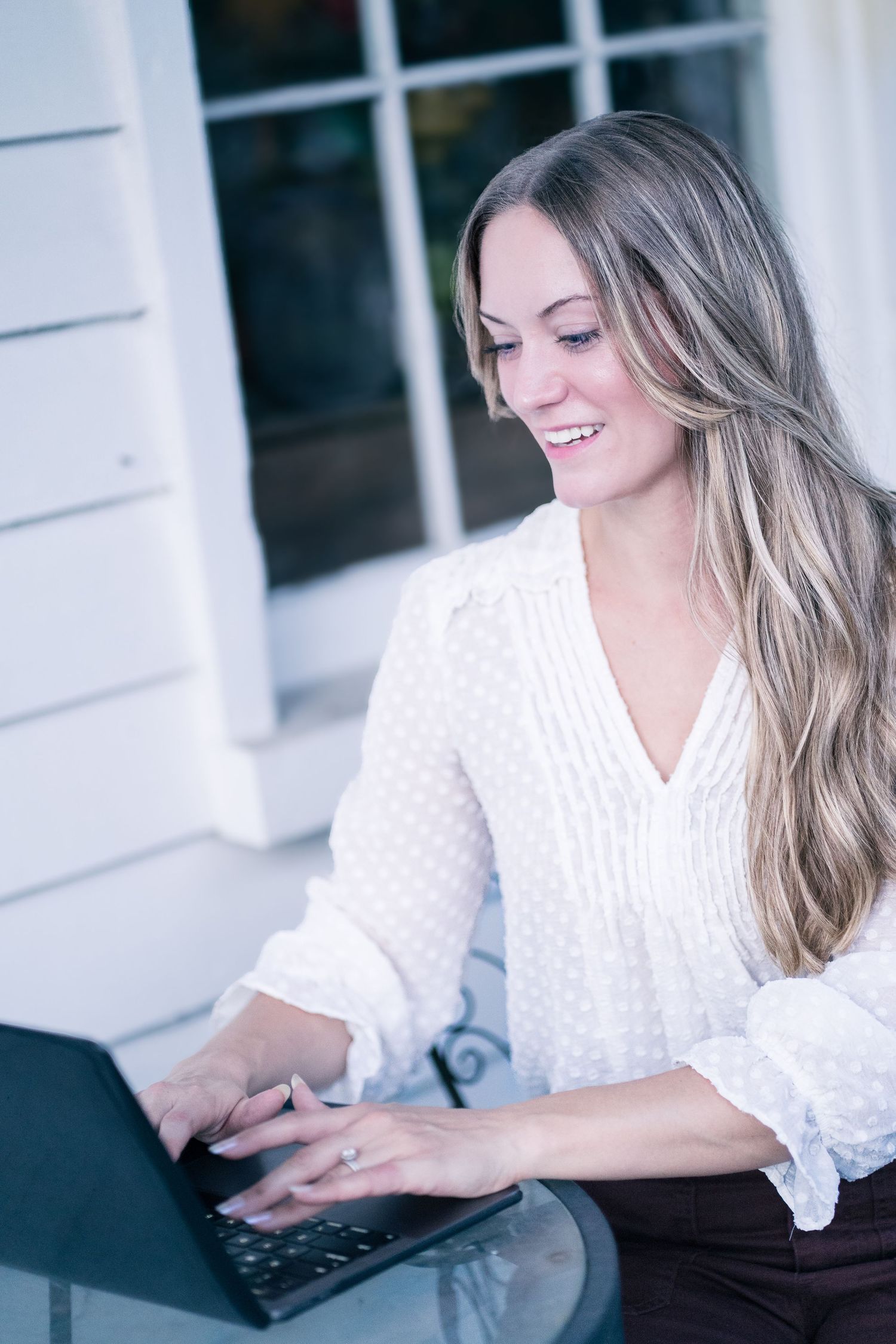 Professional with long blonde hair wearing a white blouse types on laptop while working outdoors.