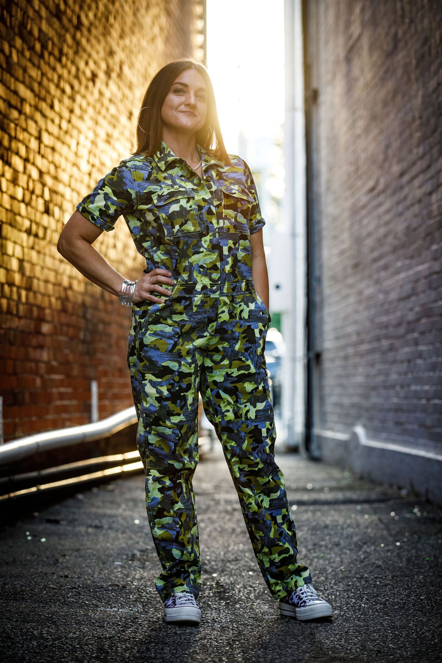 Person wearing blue camouflage pants poses against brick wall in sunset lighting creating dramatic urban silhouette.