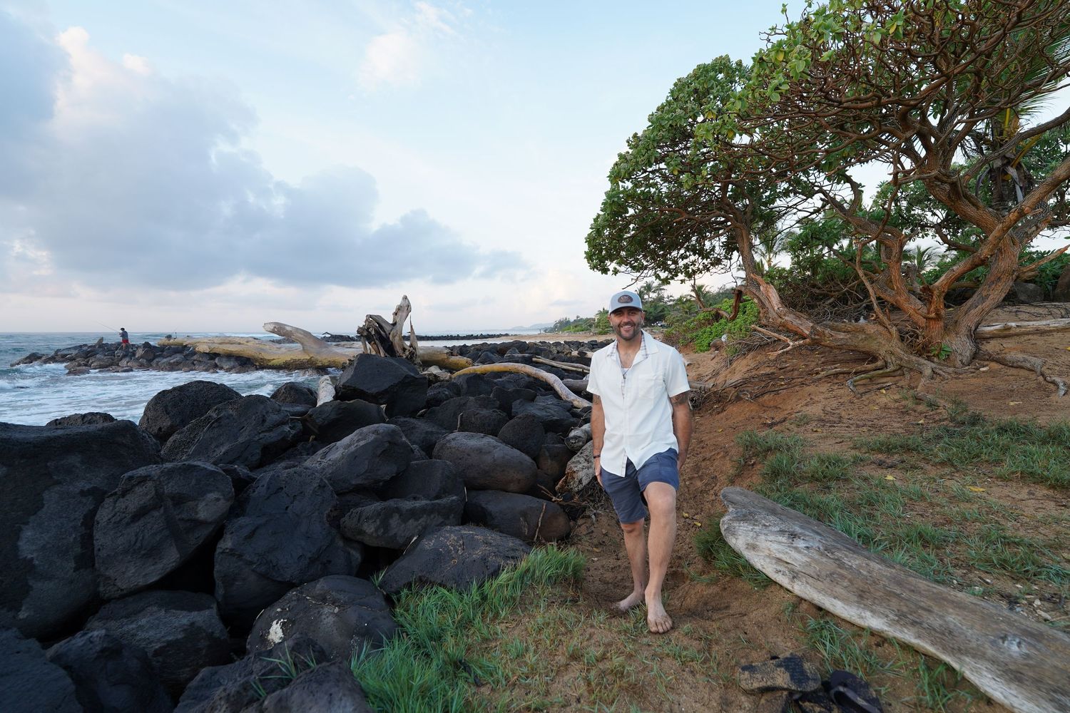 Person in white shirt standing on rocky coastline with ocean and tree in background.
