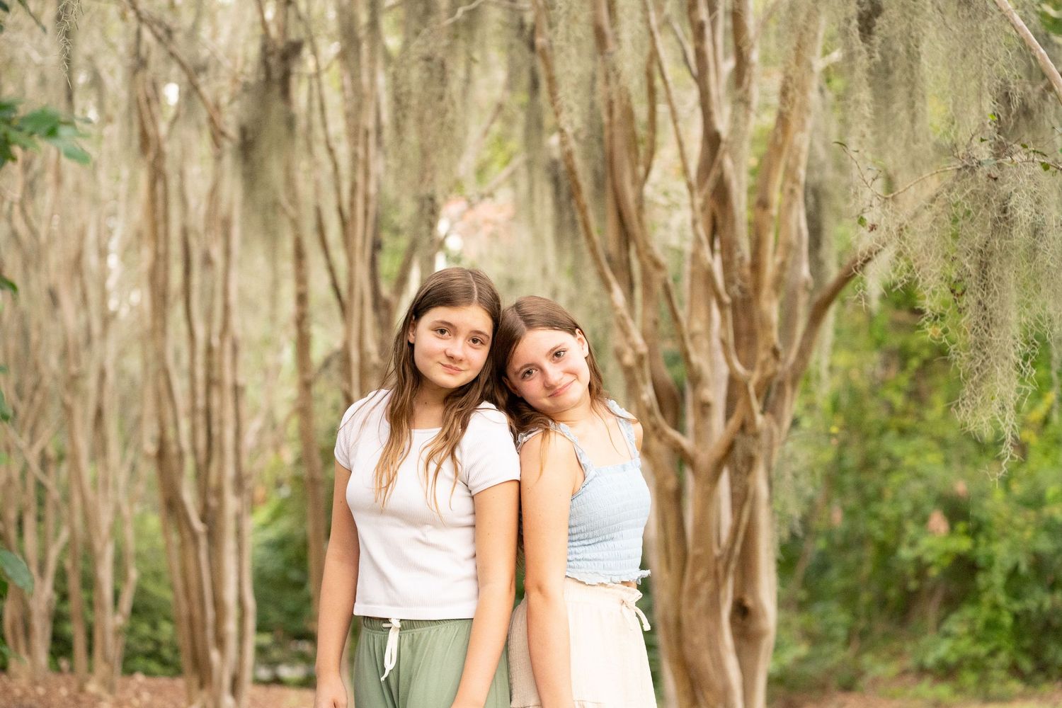 Two people in white dresses walk together through a sunlit wooded area with Spanish moss hanging from trees.