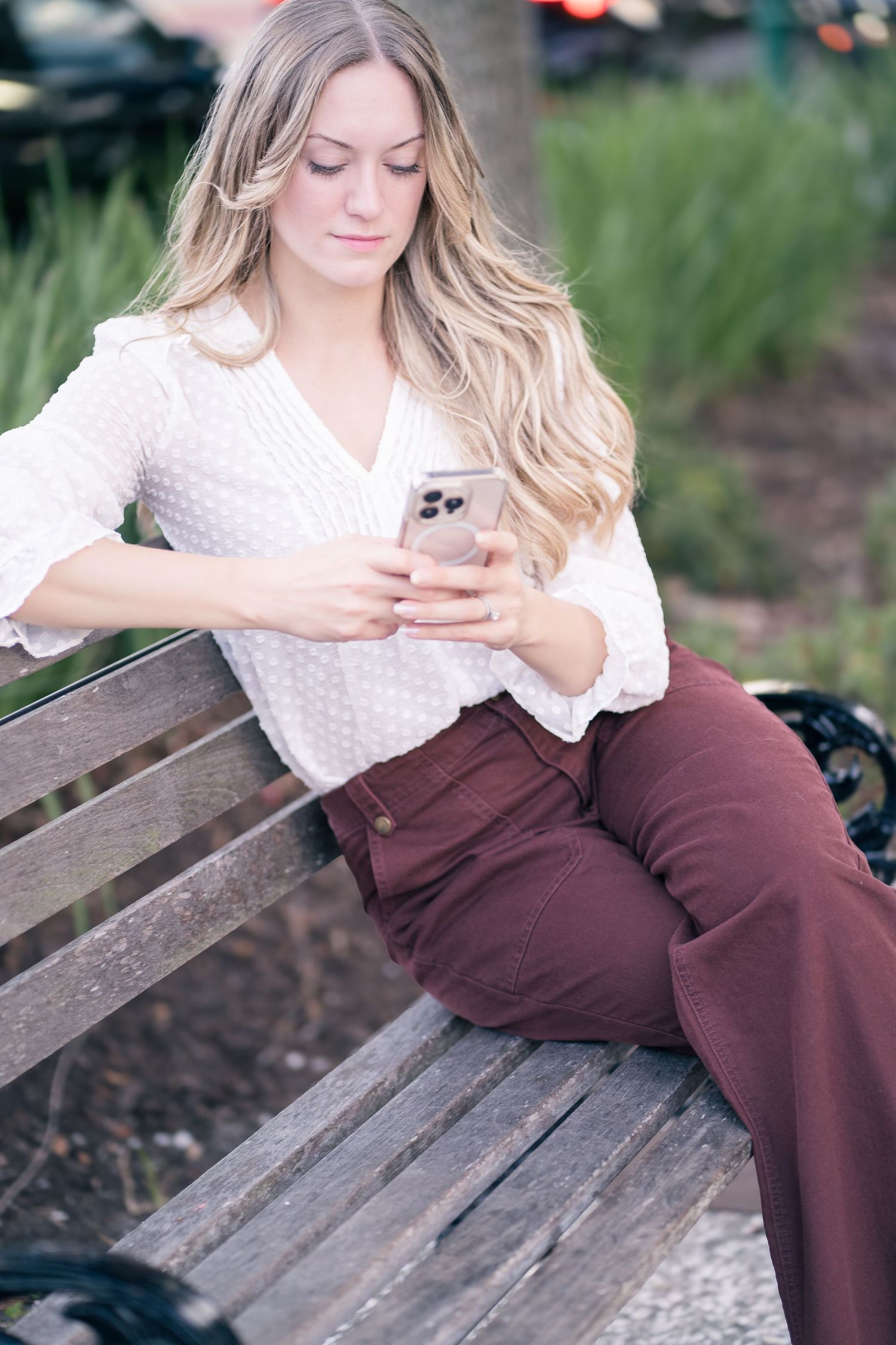 A person in a white sweater and burgundy pants sits casually on a wooden bench in an outdoor setting.