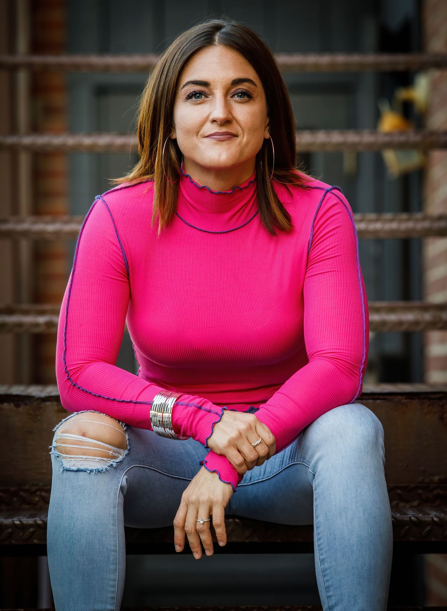 A person in a bright pink turtleneck sweater and ripped jeans sits casually on concrete steps.