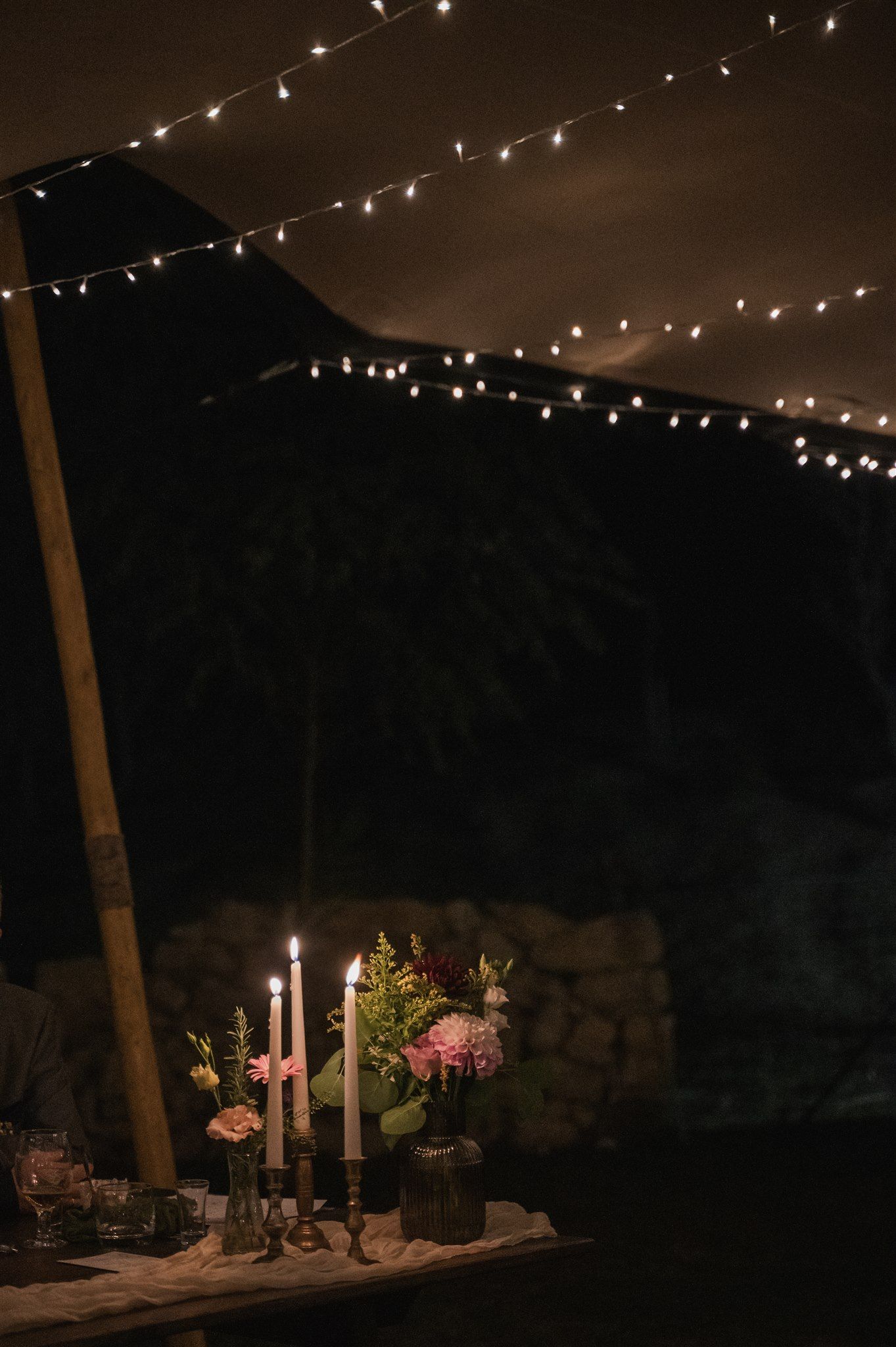 Romantic table setting with candles and flowers under string lights at night.