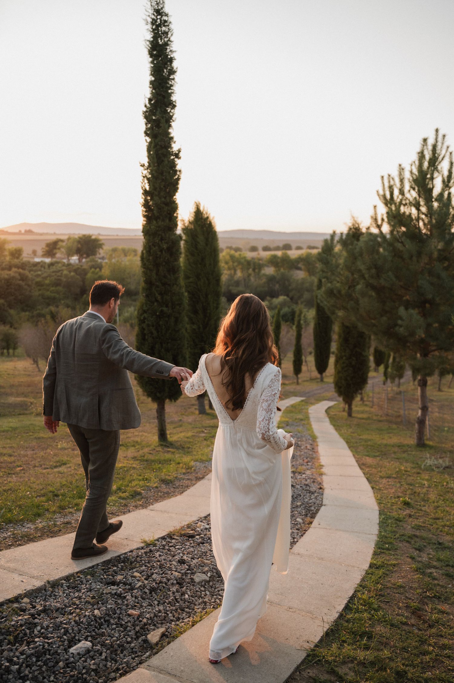 A couple walks along a winding path lined with cypress trees at sunset.