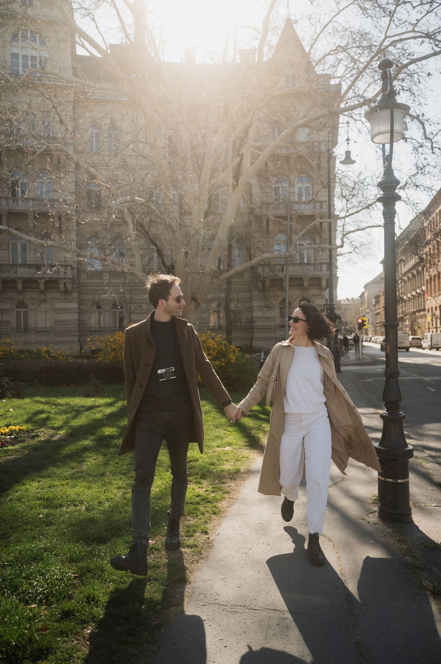 A couple walks together on a sunlit path through a city park with historic buildings in the background.