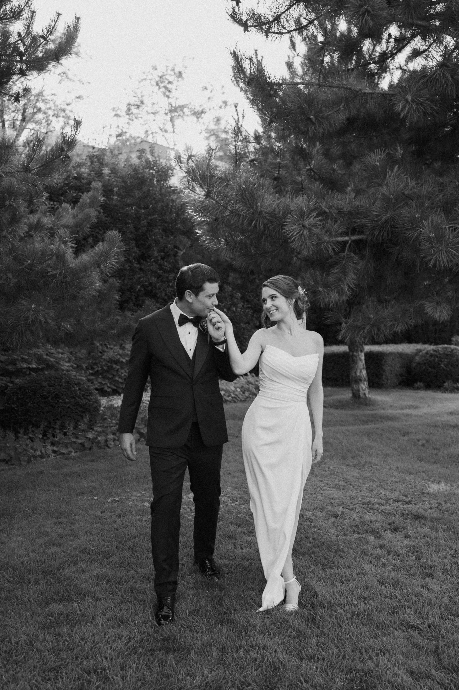 Black and white photo of newlyweds walking together on grass with trees in background.