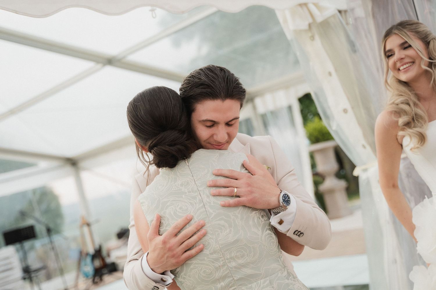 Groom shares heartfelt hug with guest while smiling bride watches at wedding reception.