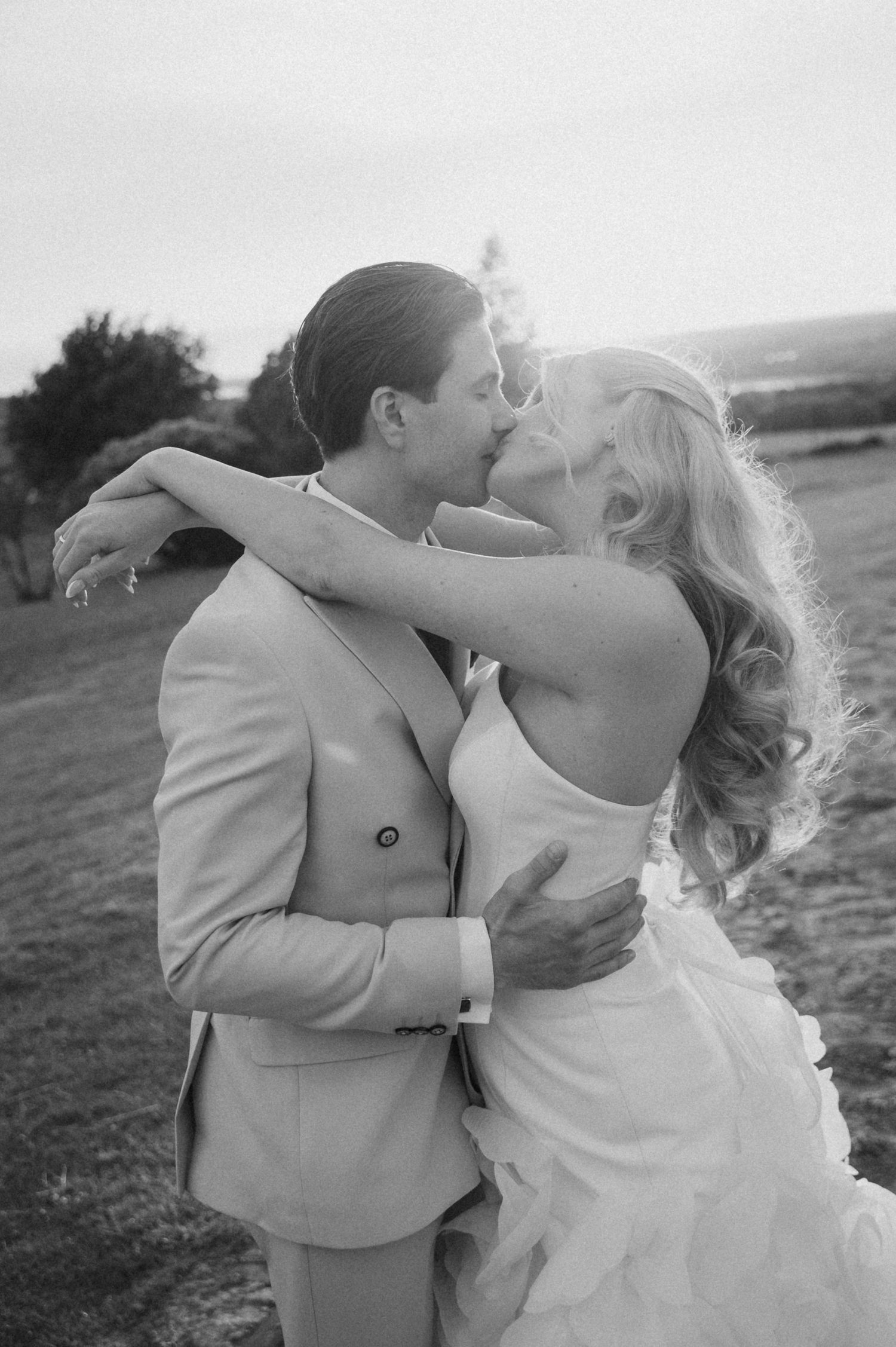 Bride and groom sharing a romantic kiss outdoors at dusk in a classic black and white wedding portrait.