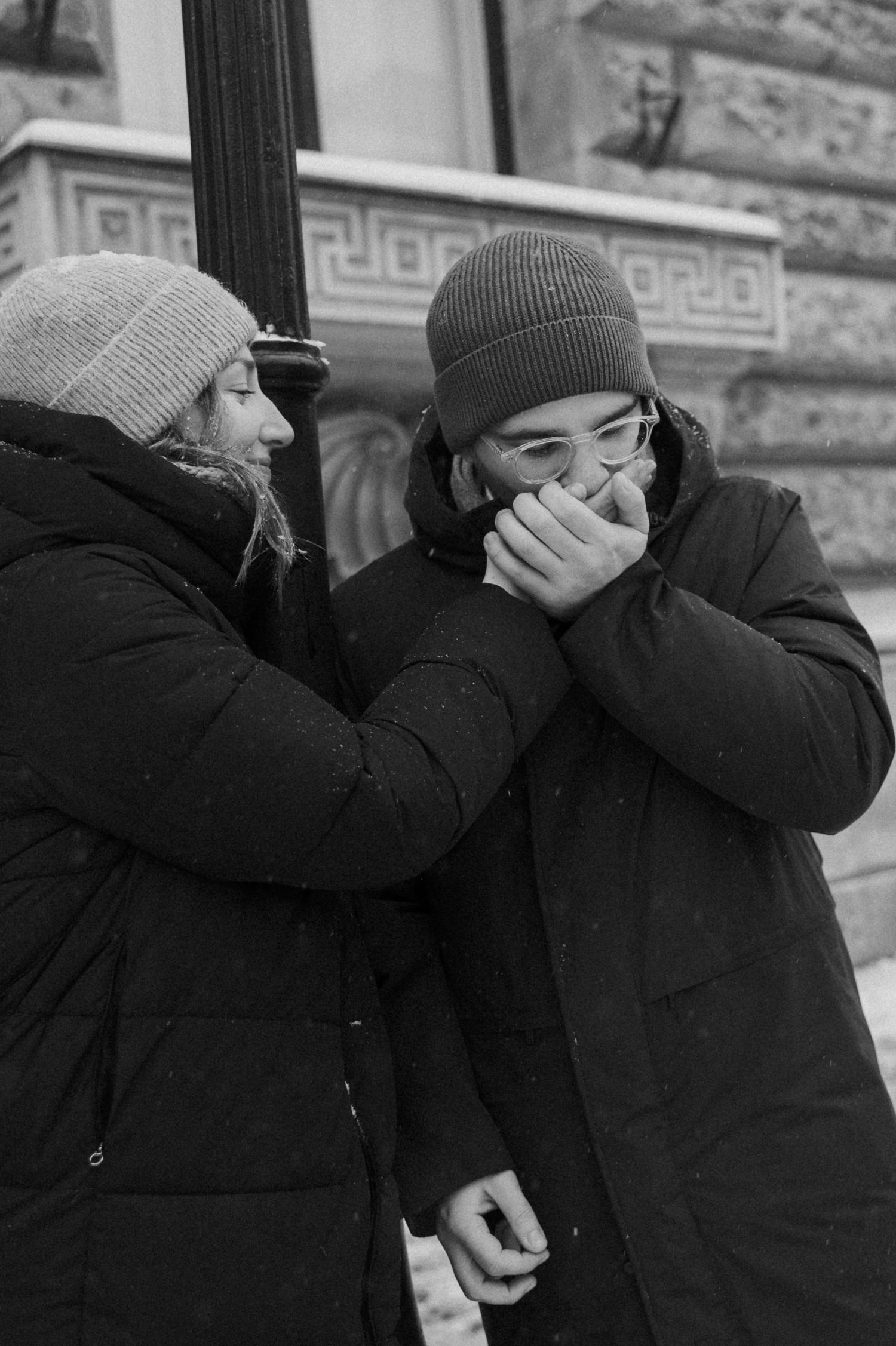 Two people share an intimate moment of laughter by a stone building in winter.
