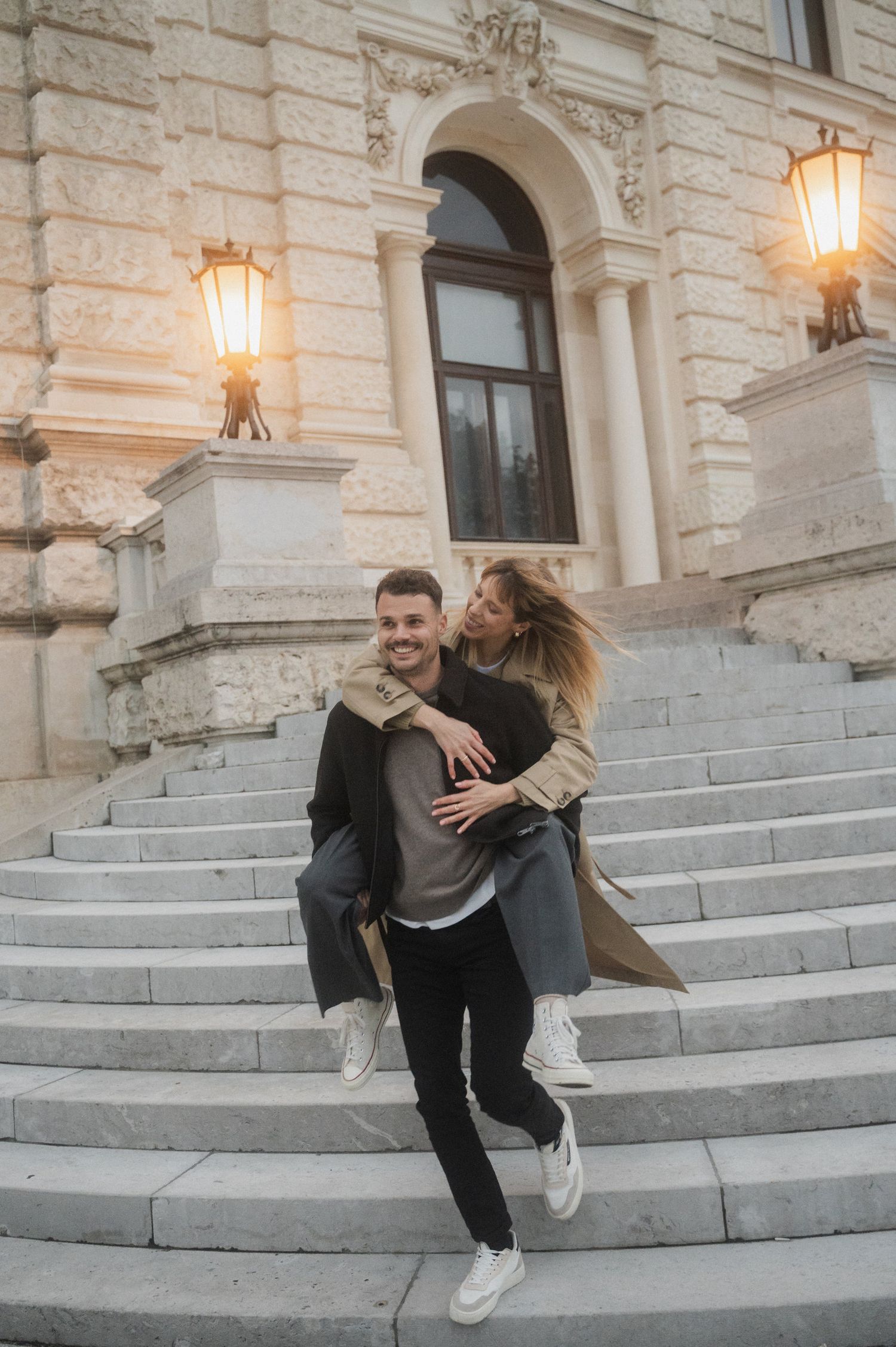 A couple shares a playful moment on grand stone steps of a historic building with illuminated lanterns at dusk.