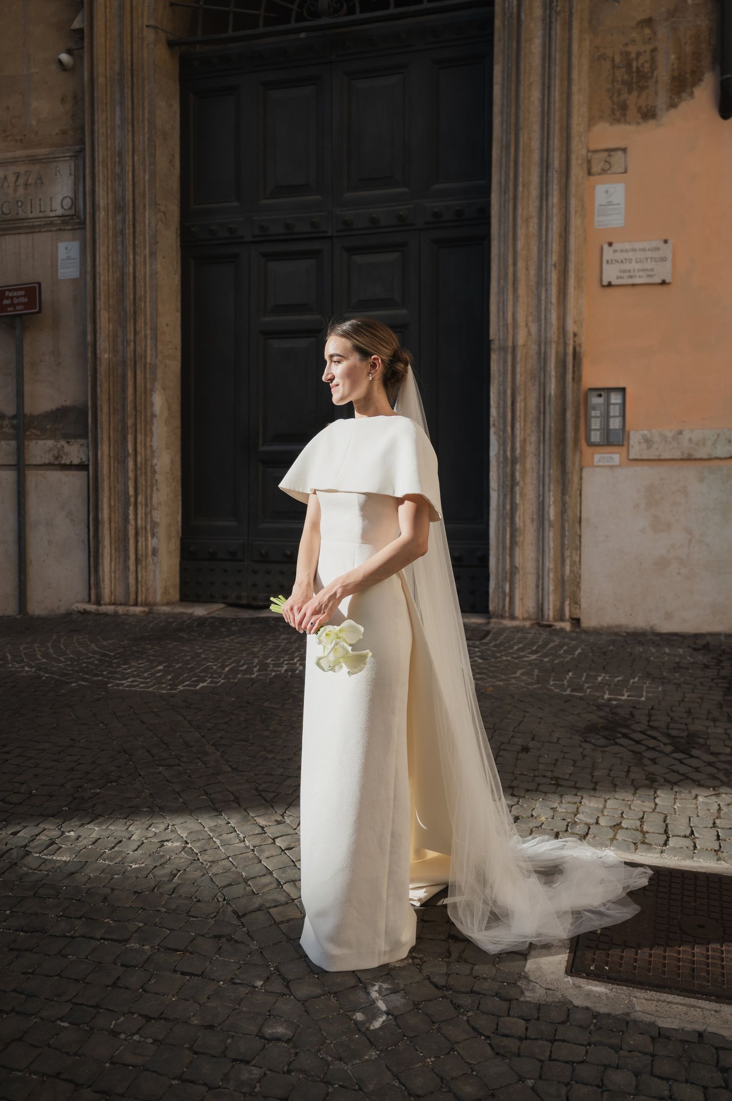 Elegant bride in white dress with flowing veil stands in historic European courtyard with cobblestone streets.