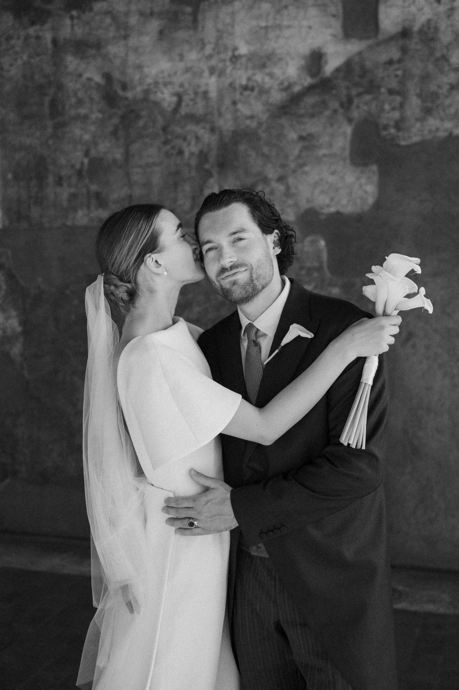 Black and white wedding portrait showing tender moment against rustic backdrop.