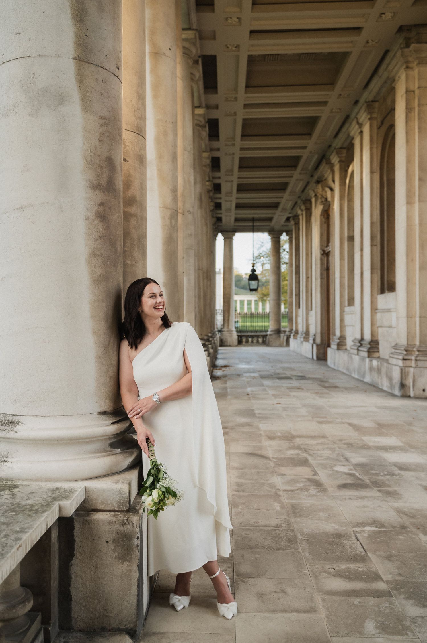 A person in a flowing white dress poses elegantly in a classical colonnade holding a bouquet.