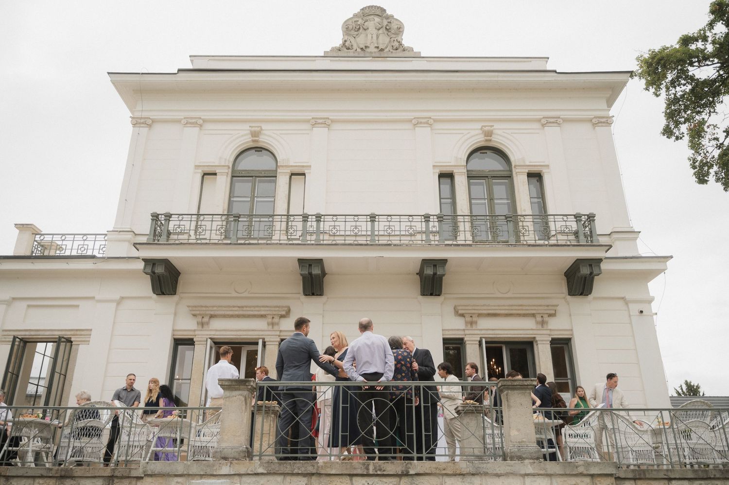 Guests gather on the terrace of an elegant white neoclassical villa during an outdoor event on a cloudy day.