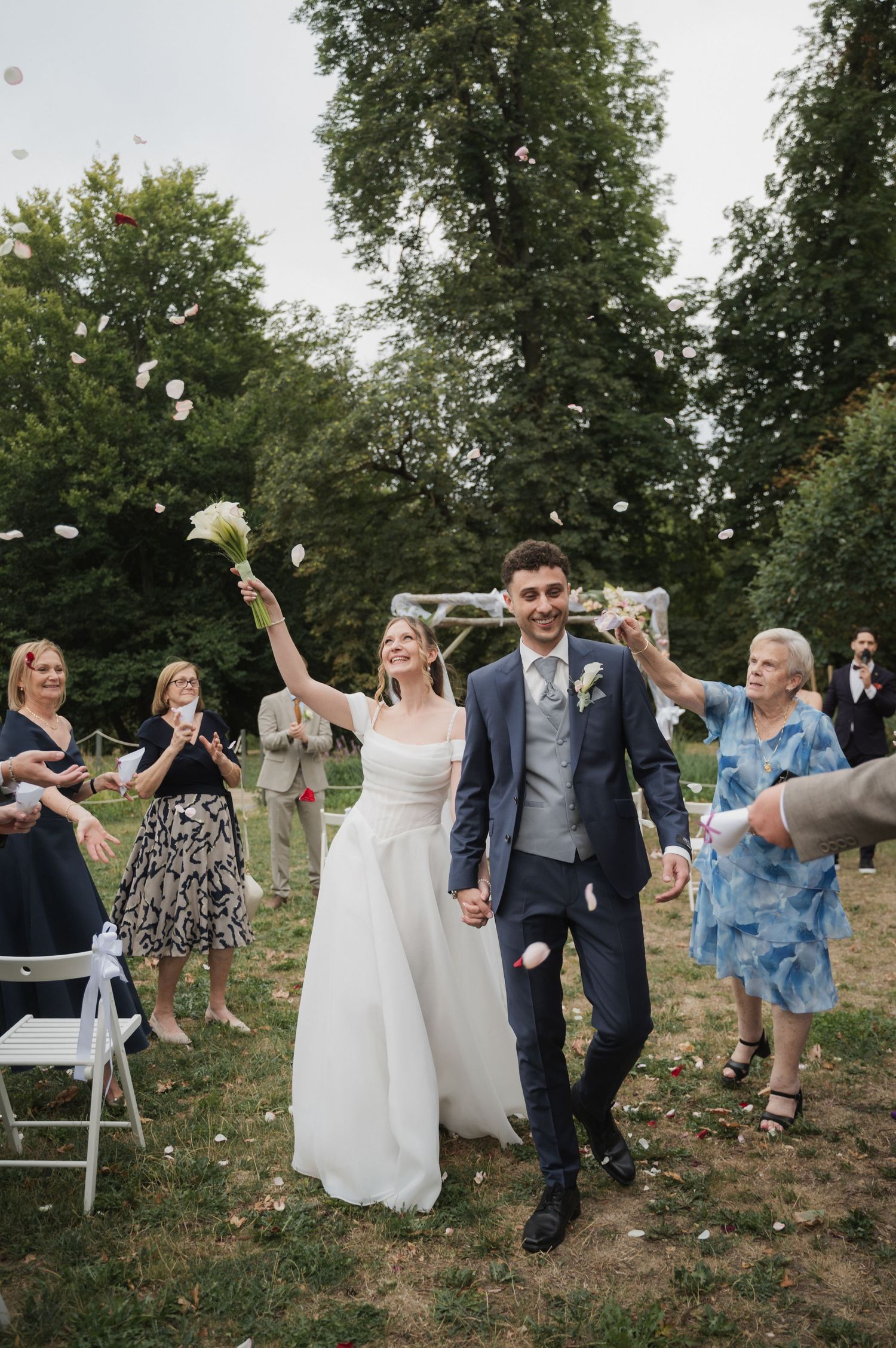 Joyful newlyweds walk through petal toss as guests celebrate outdoor wedding ceremony exit.