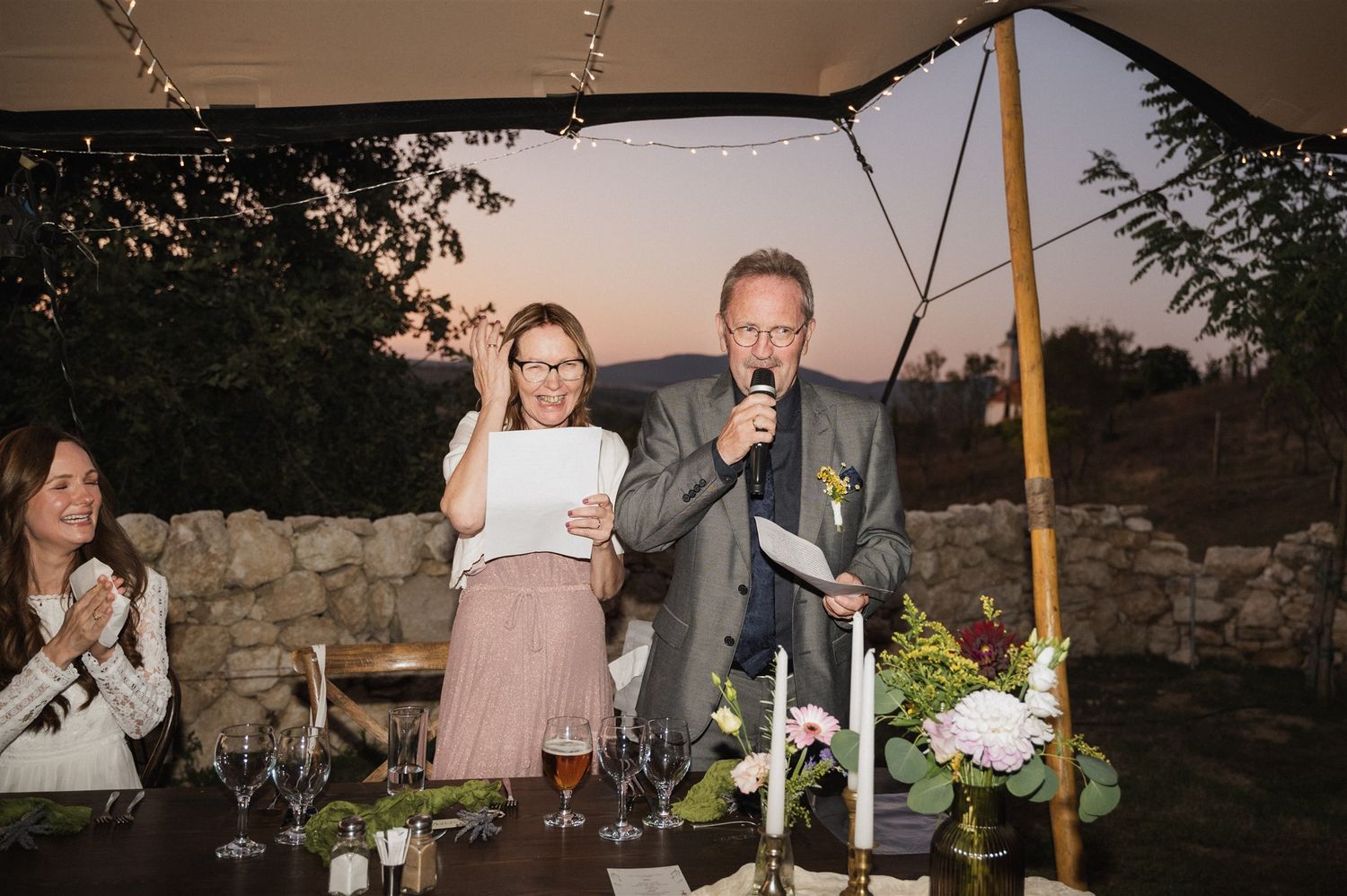 Wedding reception speech moment at sunset with stone wall backdrop and candlelit table.