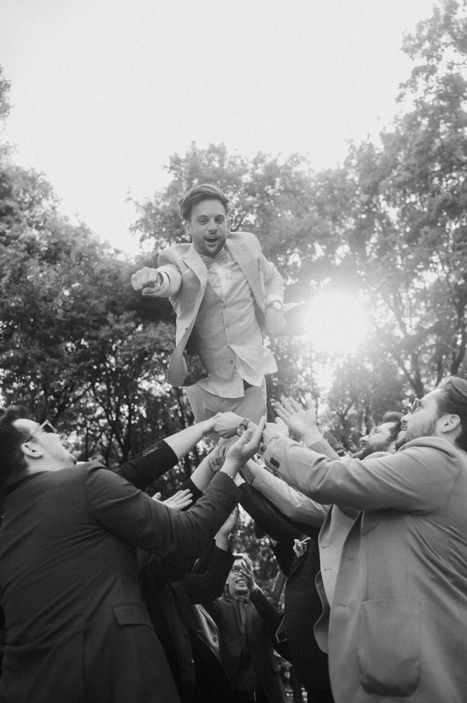 Groomsmen toss groom in air during outdoor wedding celebration in black and white photography.