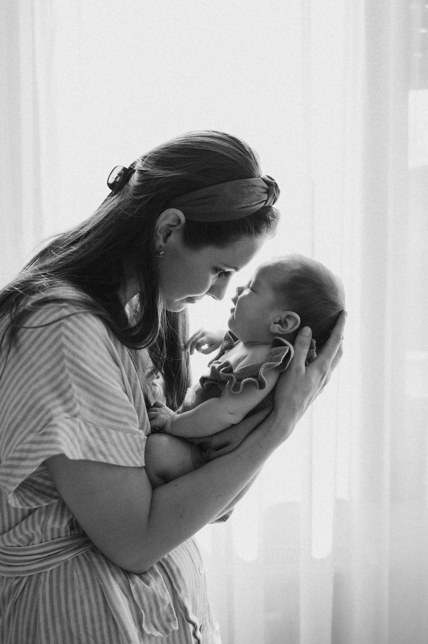 A tender black and white moment between mother and newborn baby by a sunlit window.