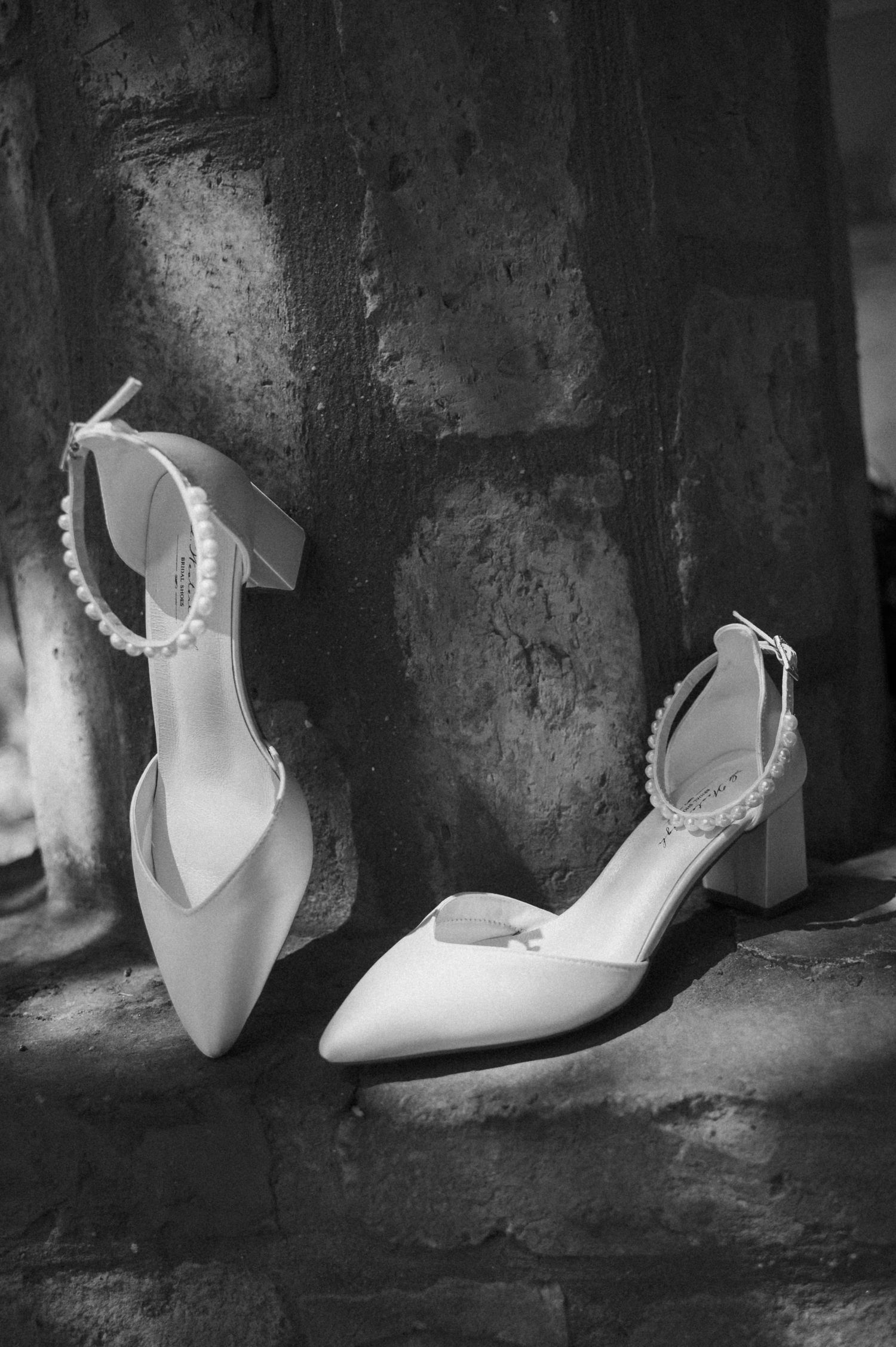 Elegant white bridal heels with pearl ankle straps displayed against a rustic stone wall in black and white.