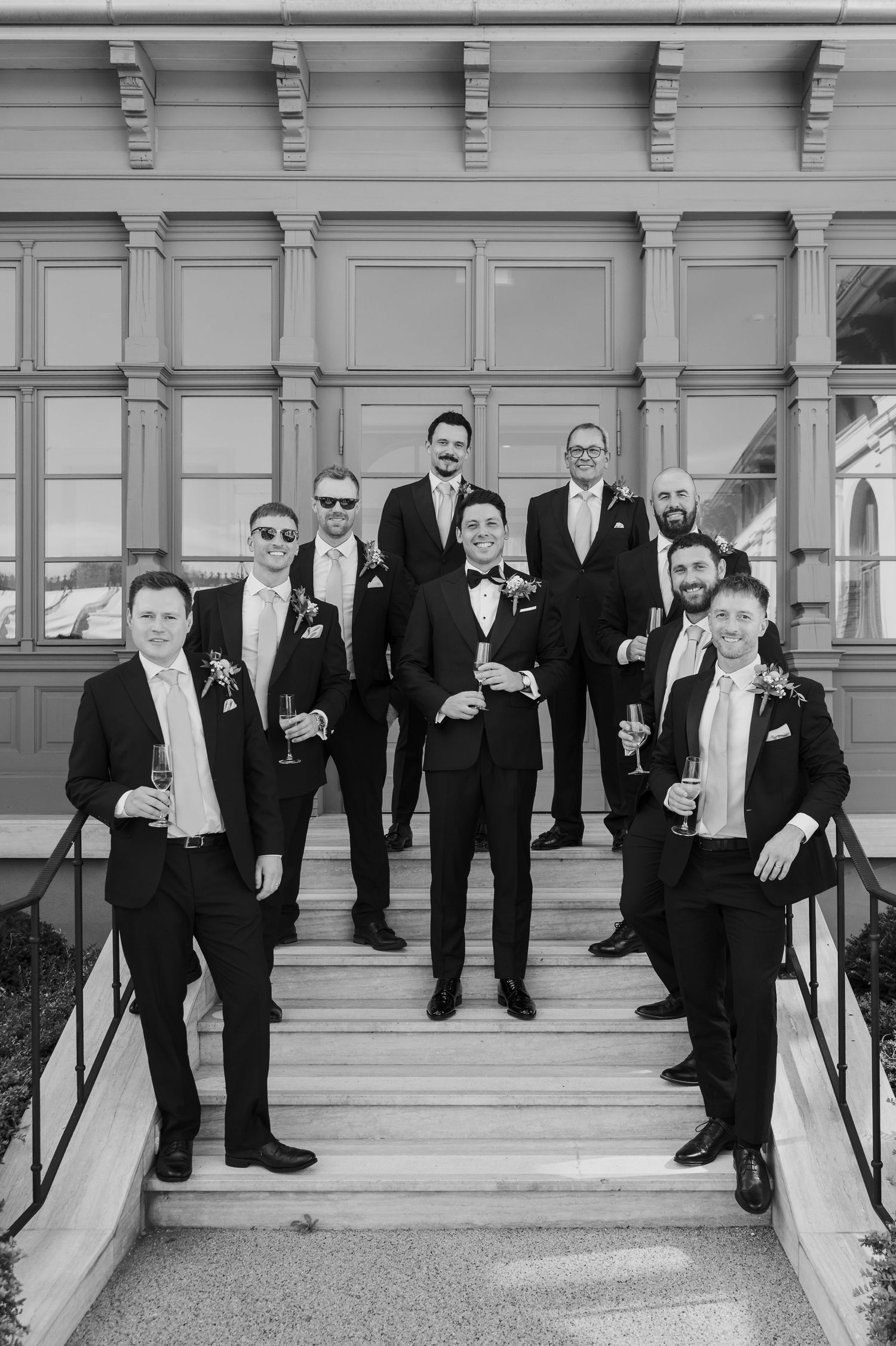 Groomsmen in black tuxedos pose on outdoor steps for formal wedding party photos in black and white.