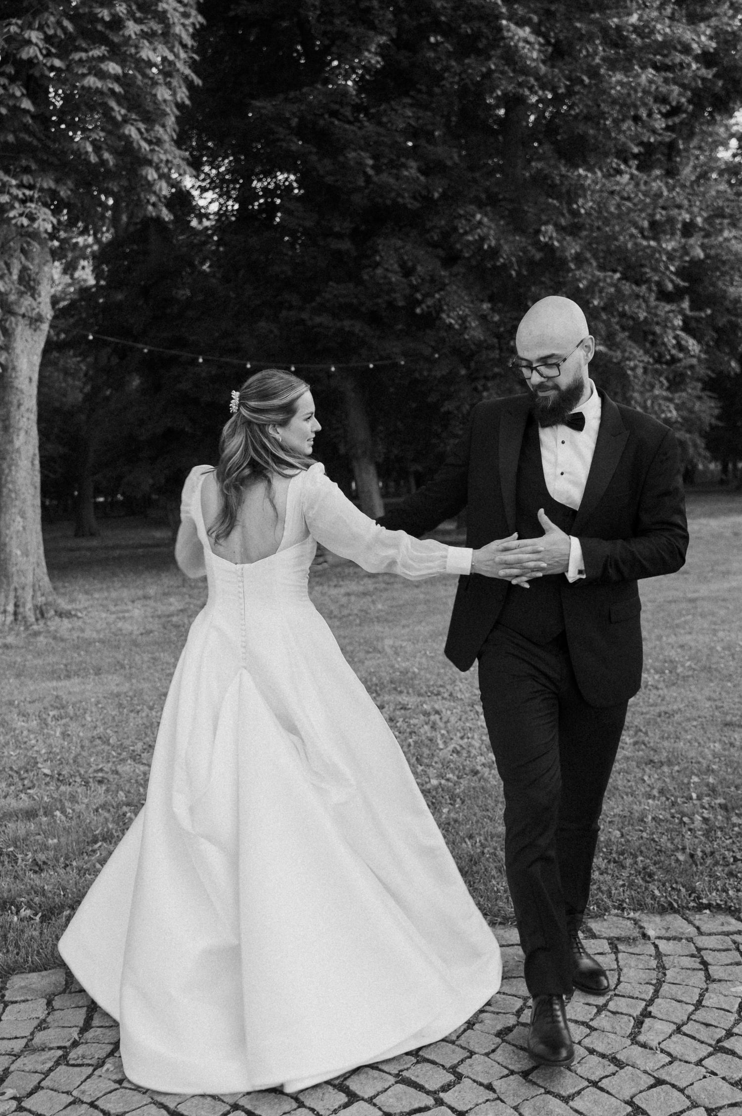 Black and white photo sequence of a couple dancing playfully on stone pavement surrounded by trees in a park setting.