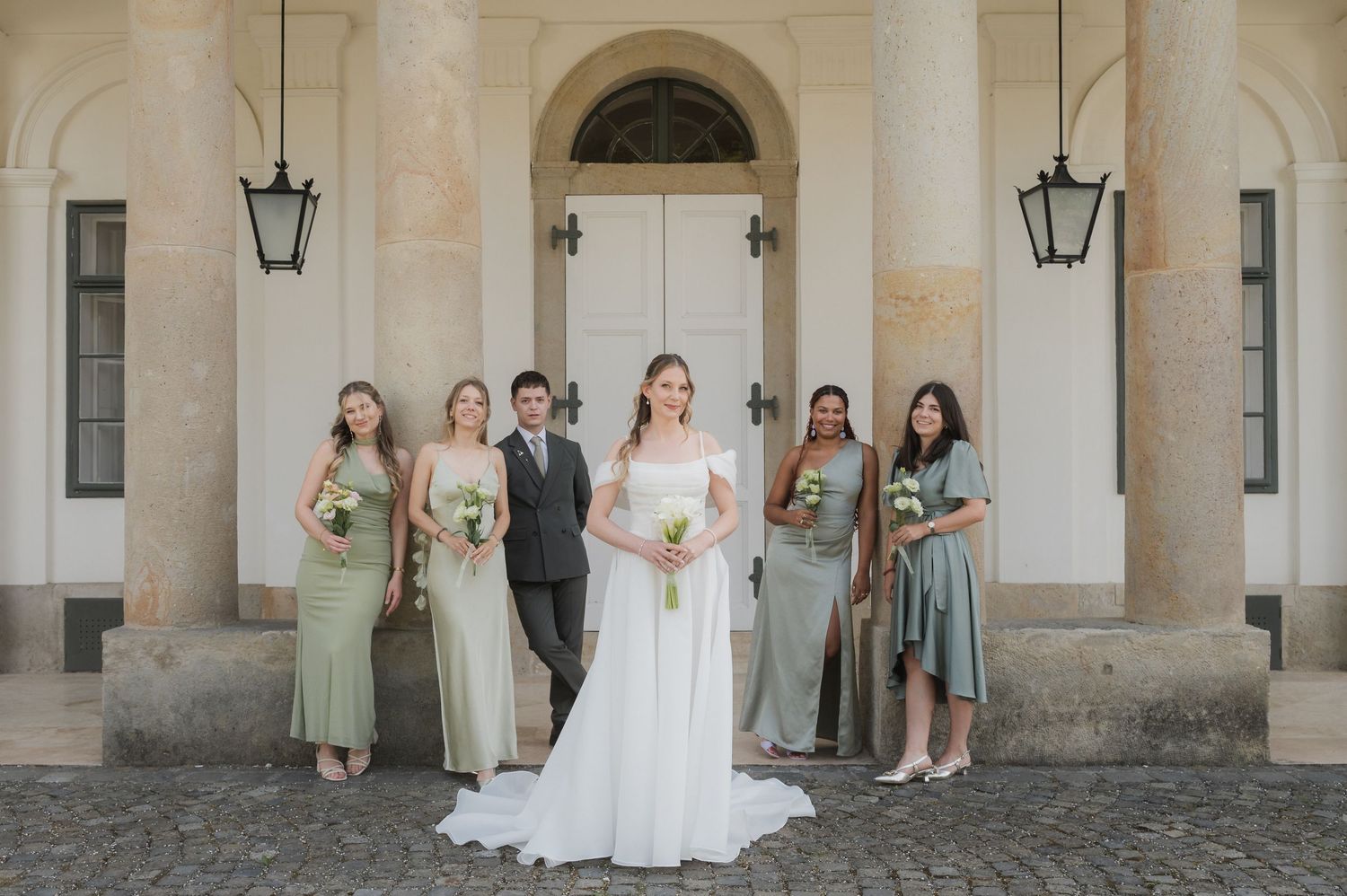 Bride in white gown poses with bridesmaids in sage green dresses before elegant white columned building.