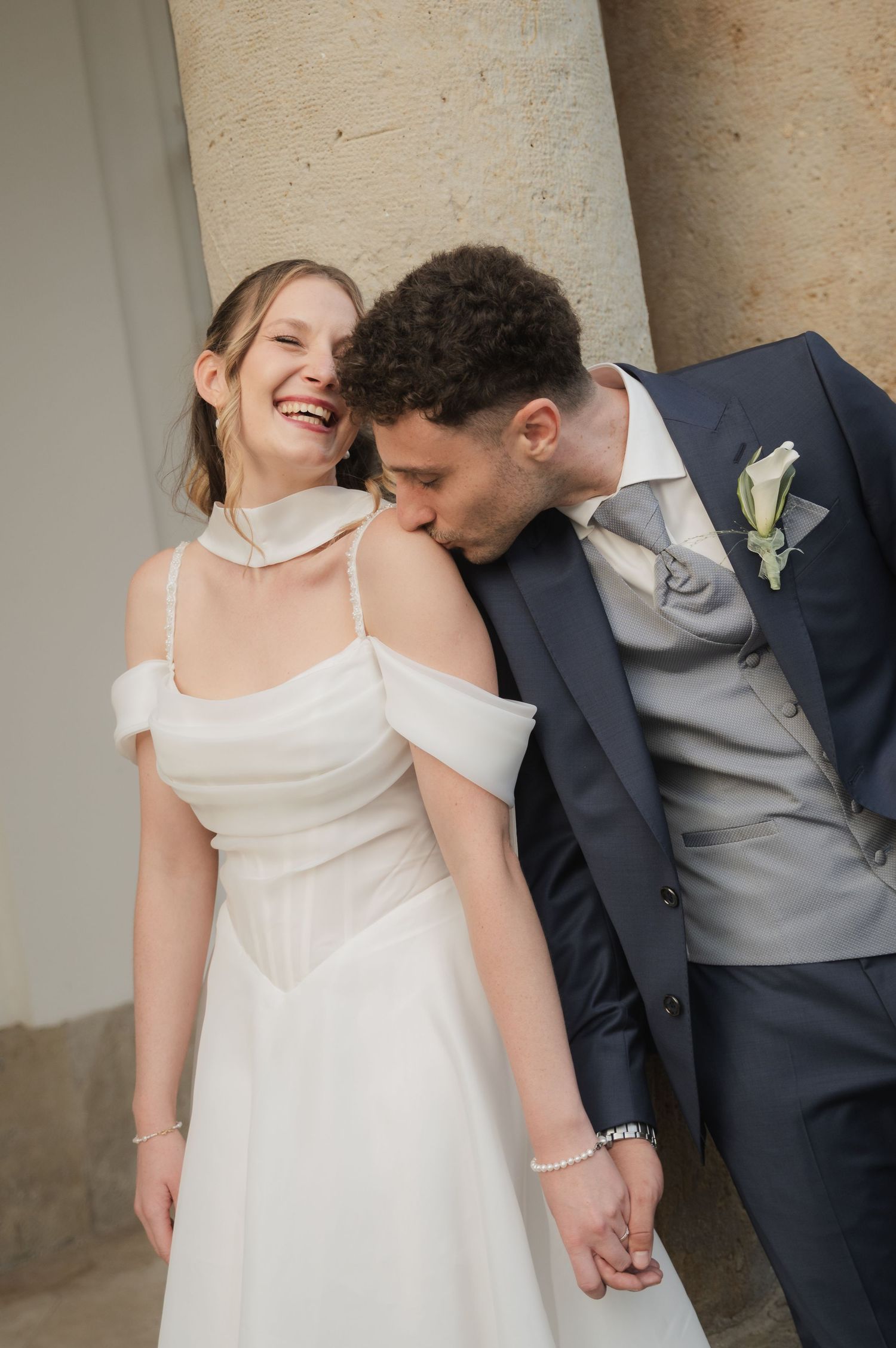 A groom in a navy suit kisses his laughing bride on the shoulder as she wears a white off-shoulder wedding dress.