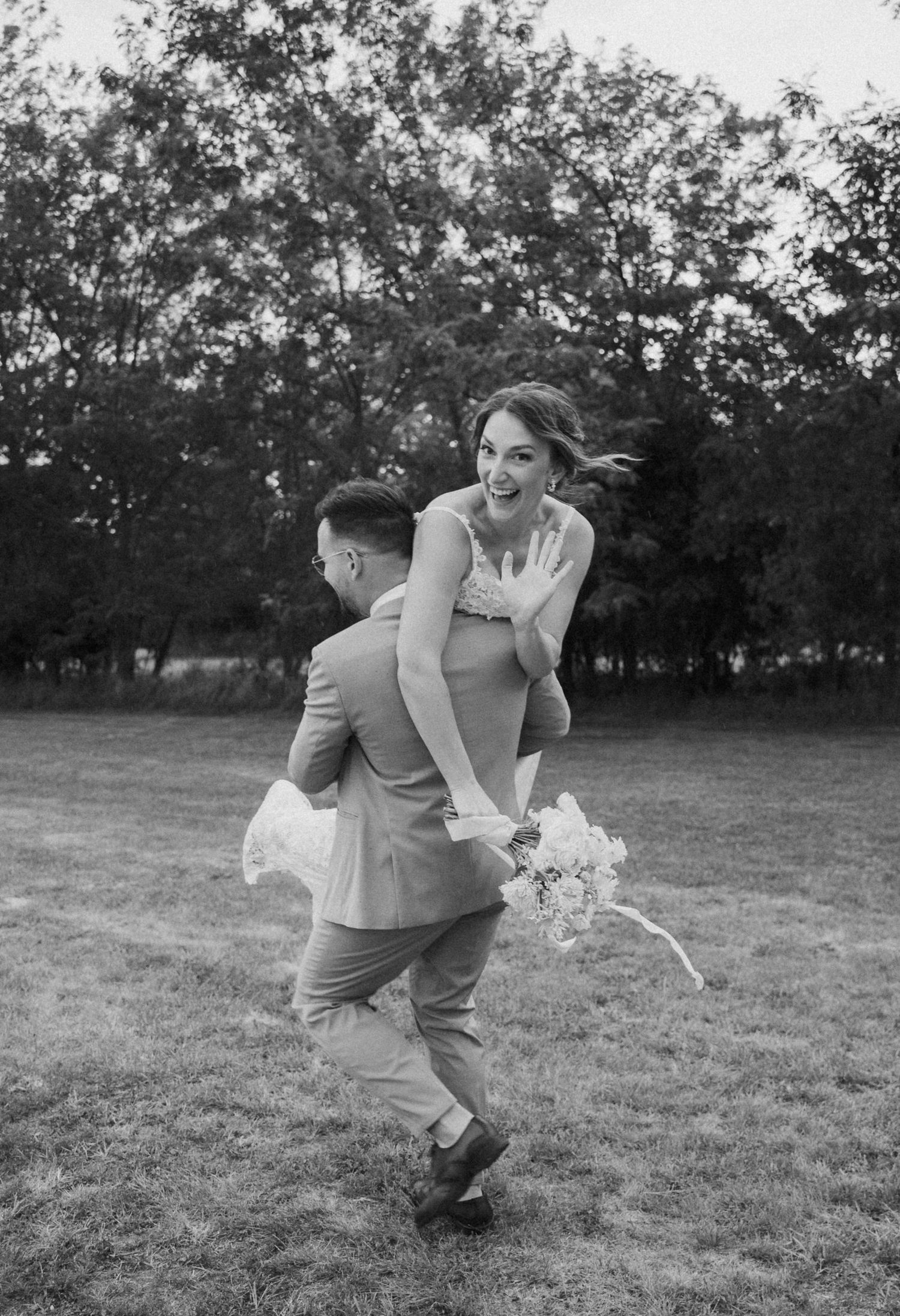 Playful wedding couple dances and spins in a grassy field at sunset.