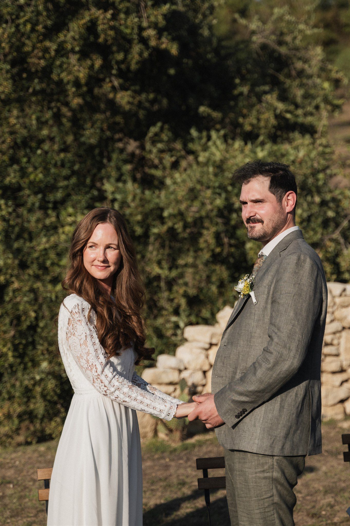 A couple holds hands during their outdoor ceremony with stone wall and greenery backdrop.