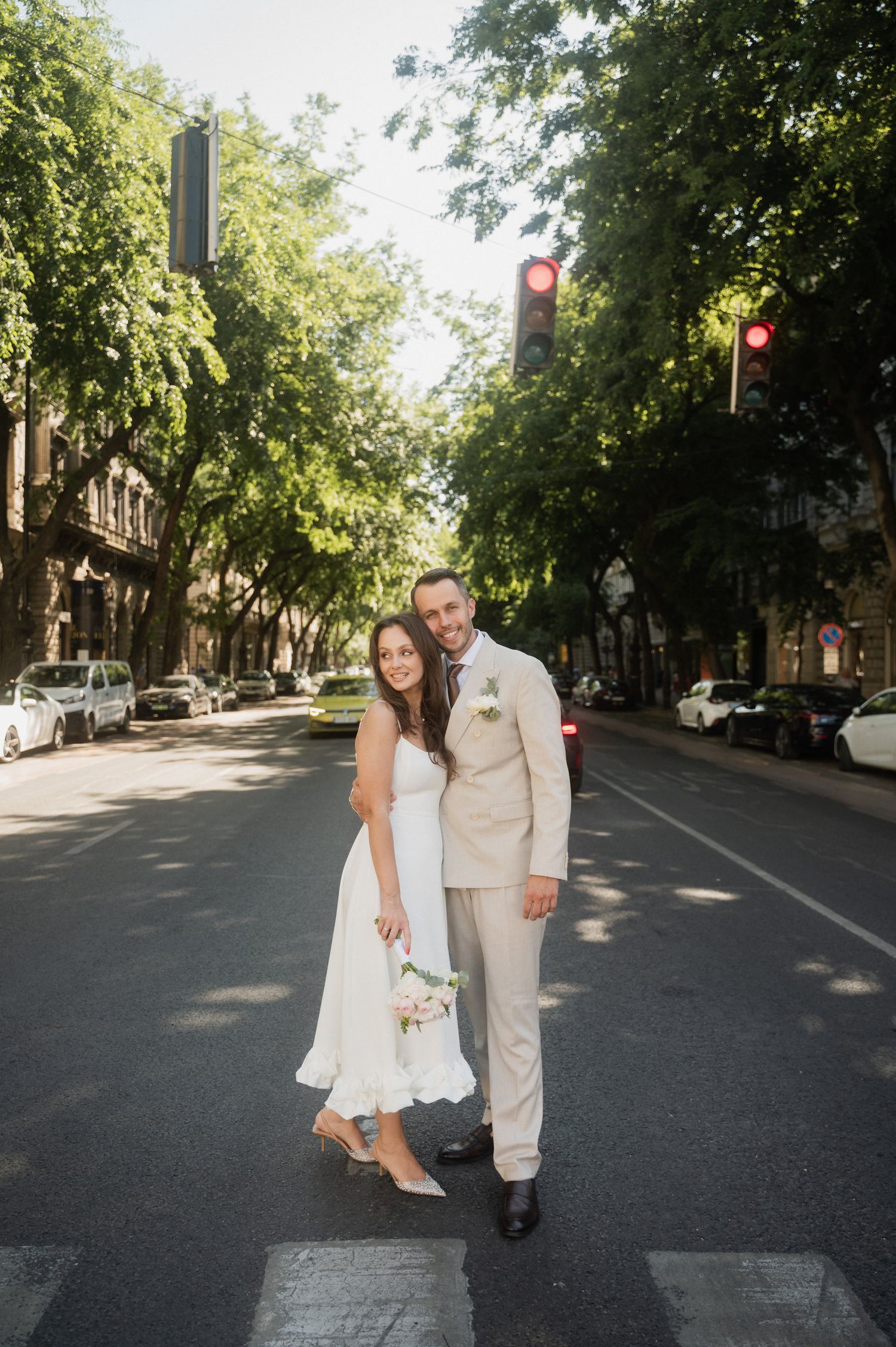 Couple in wedding attire dances playfully on tree-lined street.
