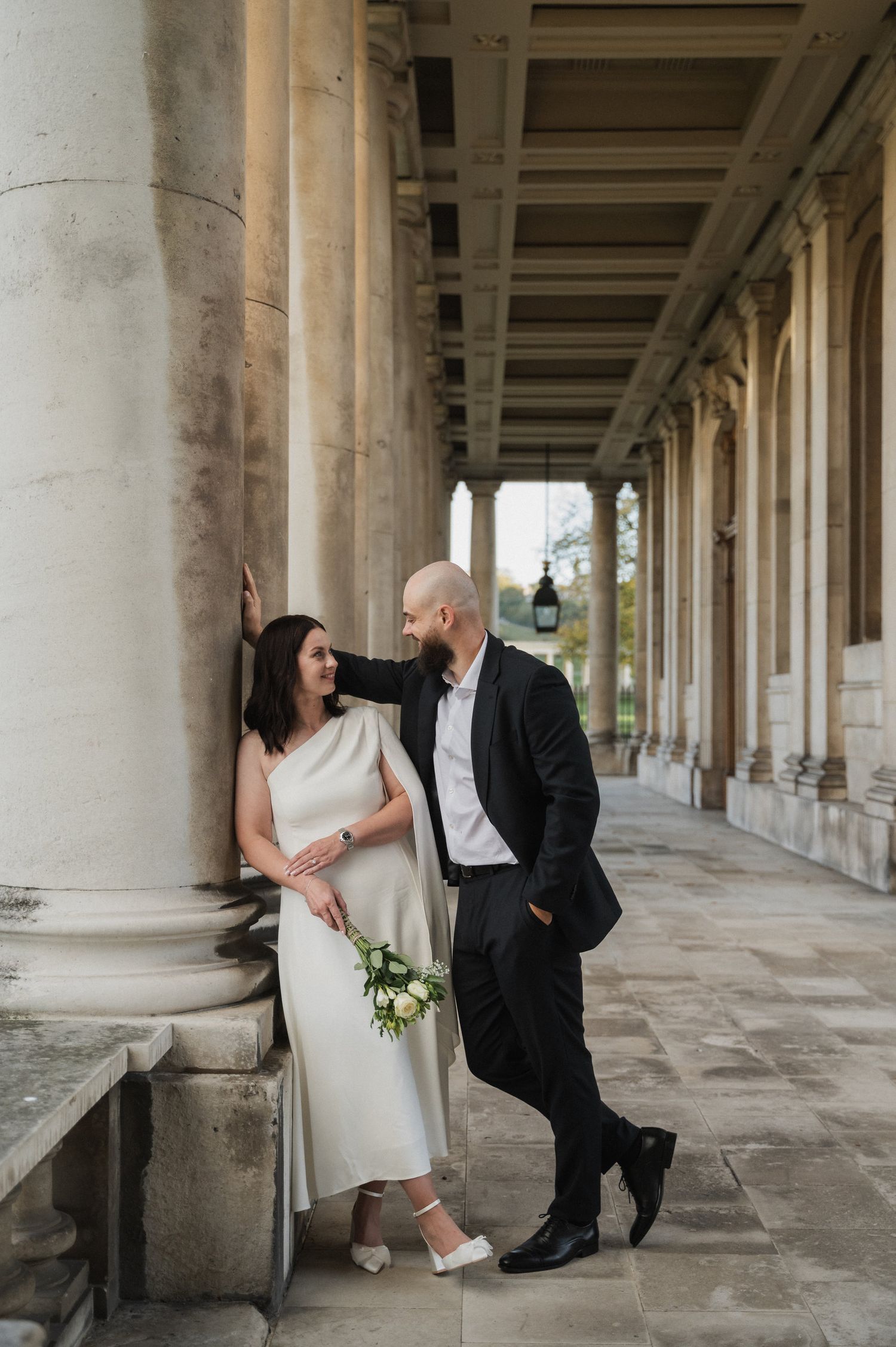 A couple in wedding attire shares an intimate moment among classical stone columns.
