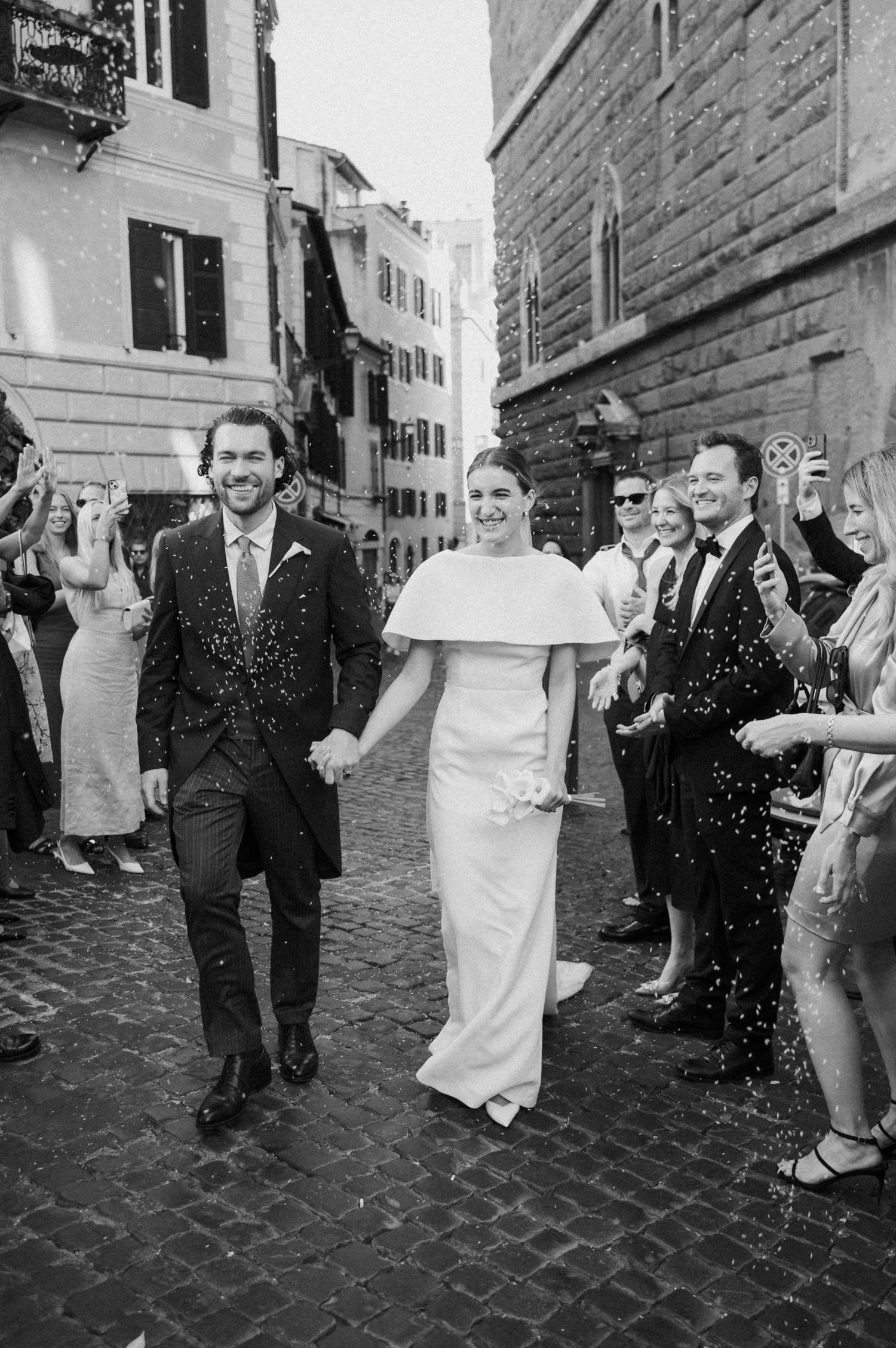 Black and white photo of wedding party walking down cobblestone street during celebration.