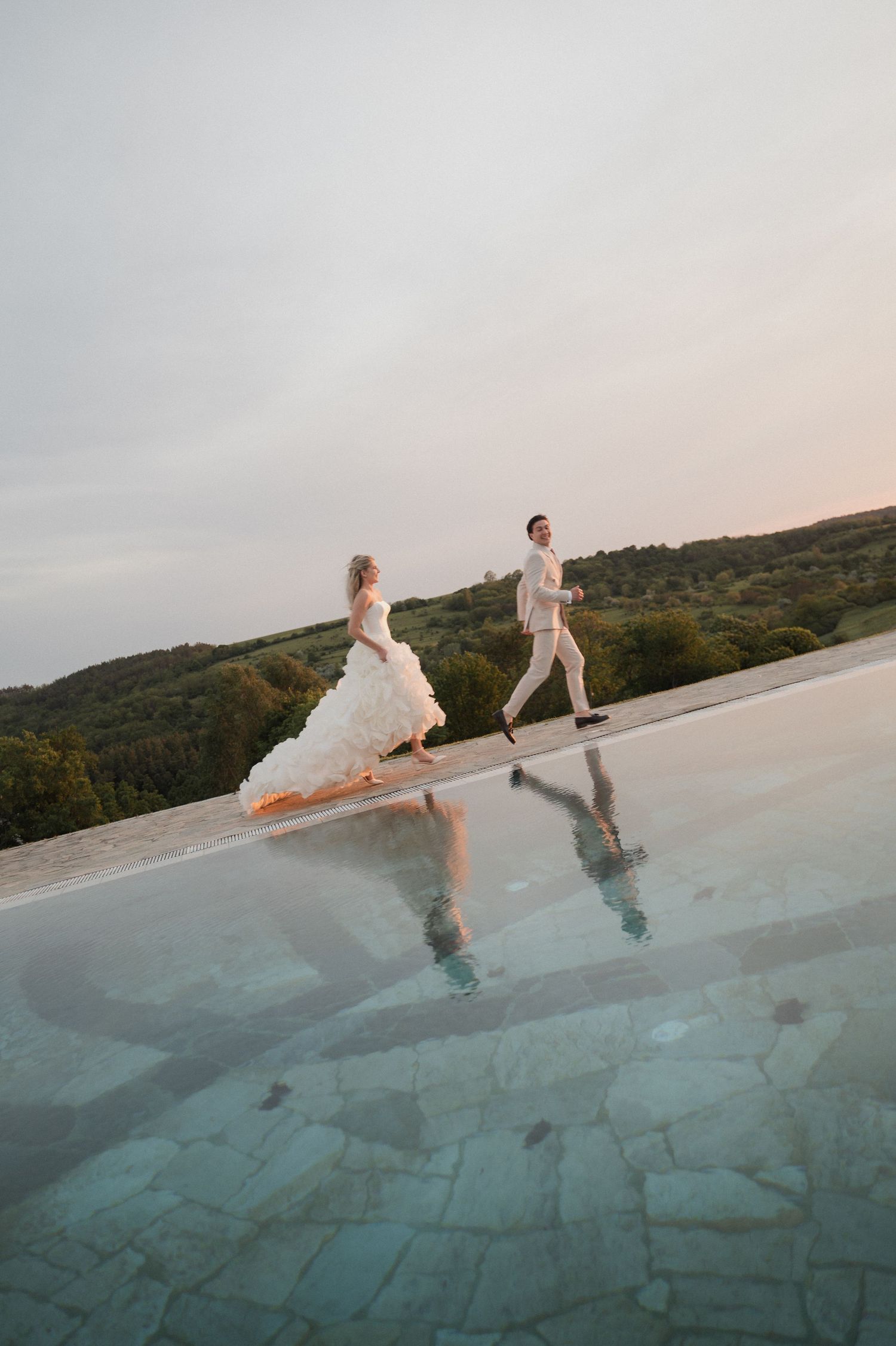 Bride and groom run joyfully beside infinity pool at sunset with scenic green hills backdrop.