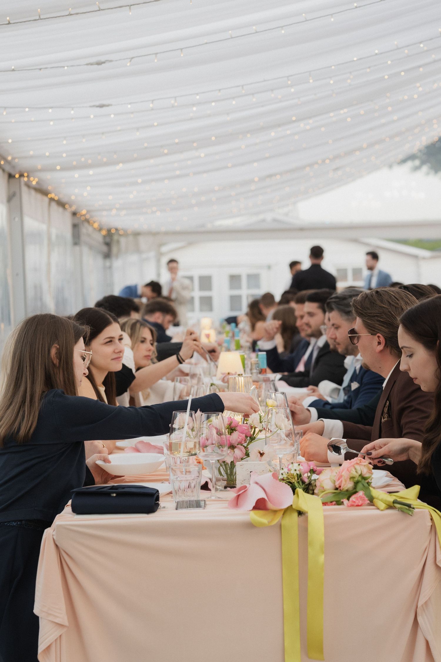 Wedding guests clink glasses at a beautifully decorated reception table under a tent with string lights.
