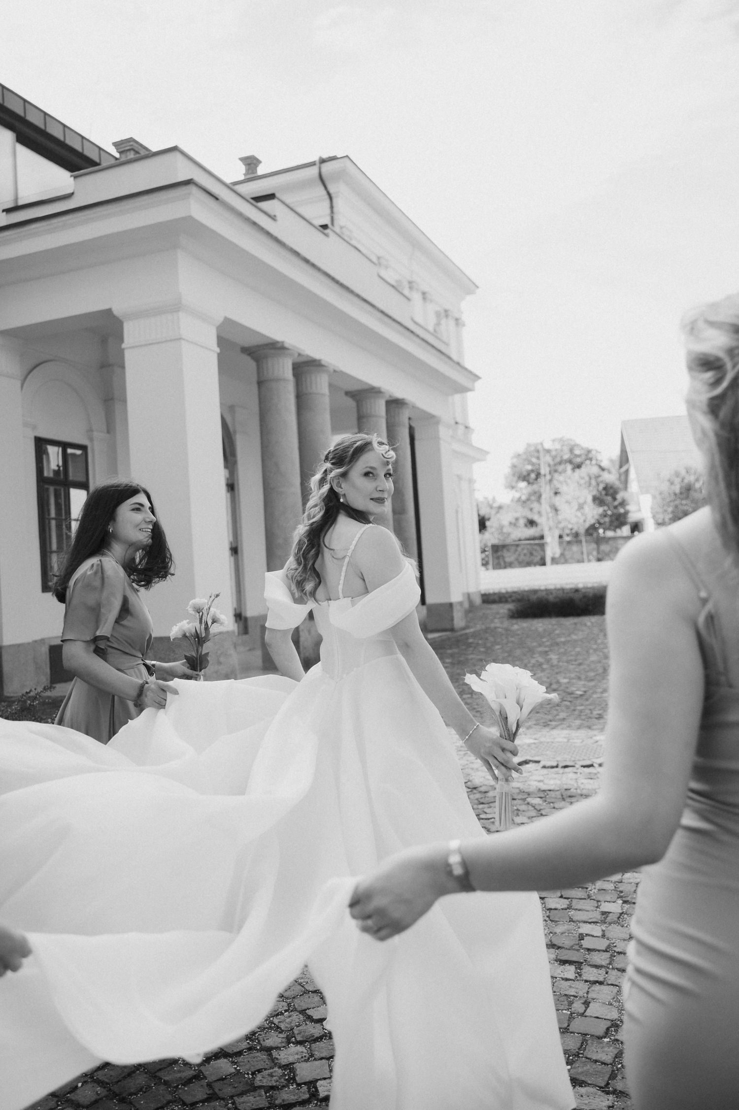 Black and white photo of a bride in a flowing gown with bridesmaids outside an elegant building.