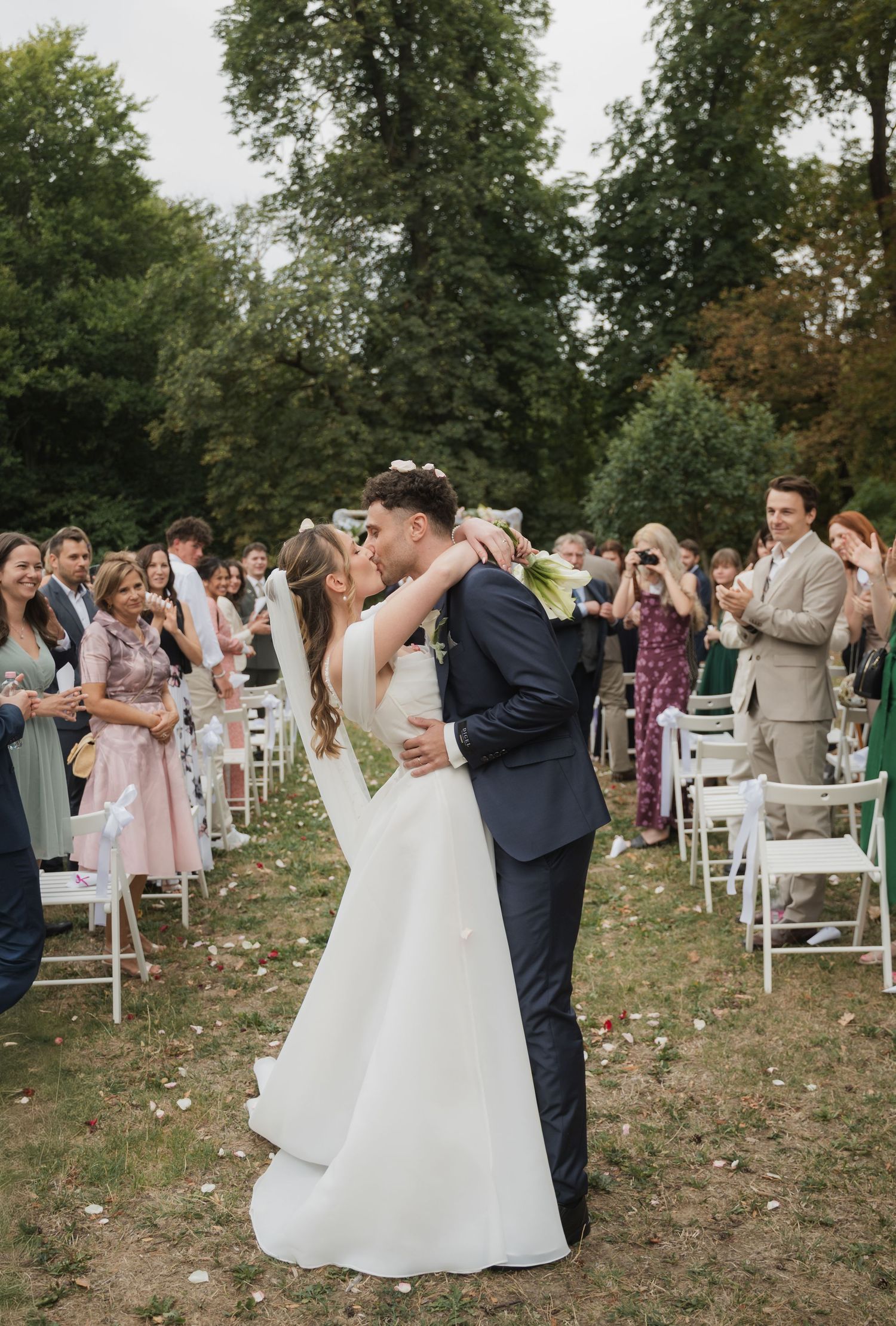 Bride and groom share a first kiss outdoors as wedding guests celebrate along the aisle.