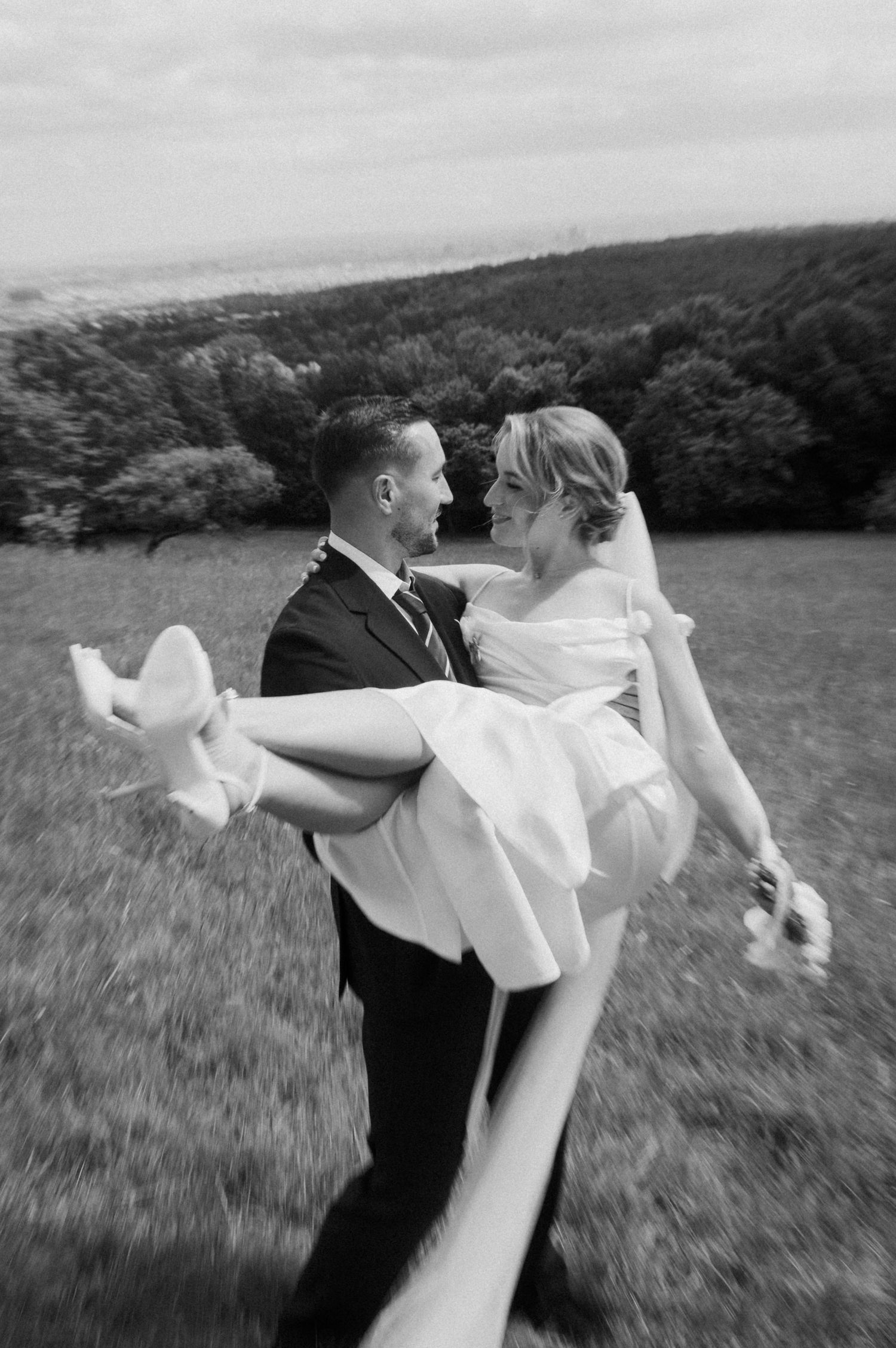 Groom carries bride in white dress across green hillside in romantic black and white wedding photo.