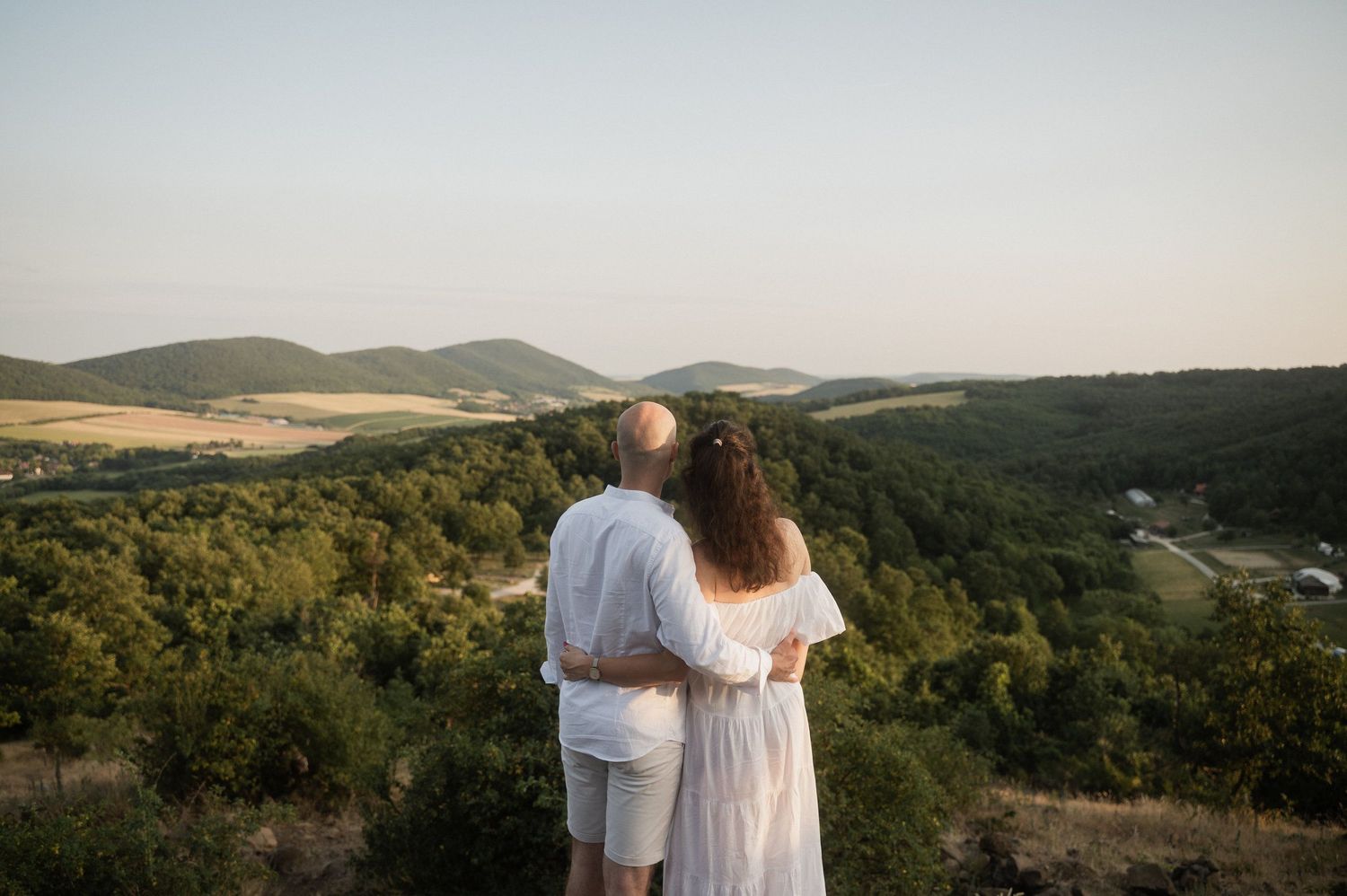 A couple in white clothing stands on a hilltop overlooking a scenic valley with rolling green hills at sunset.