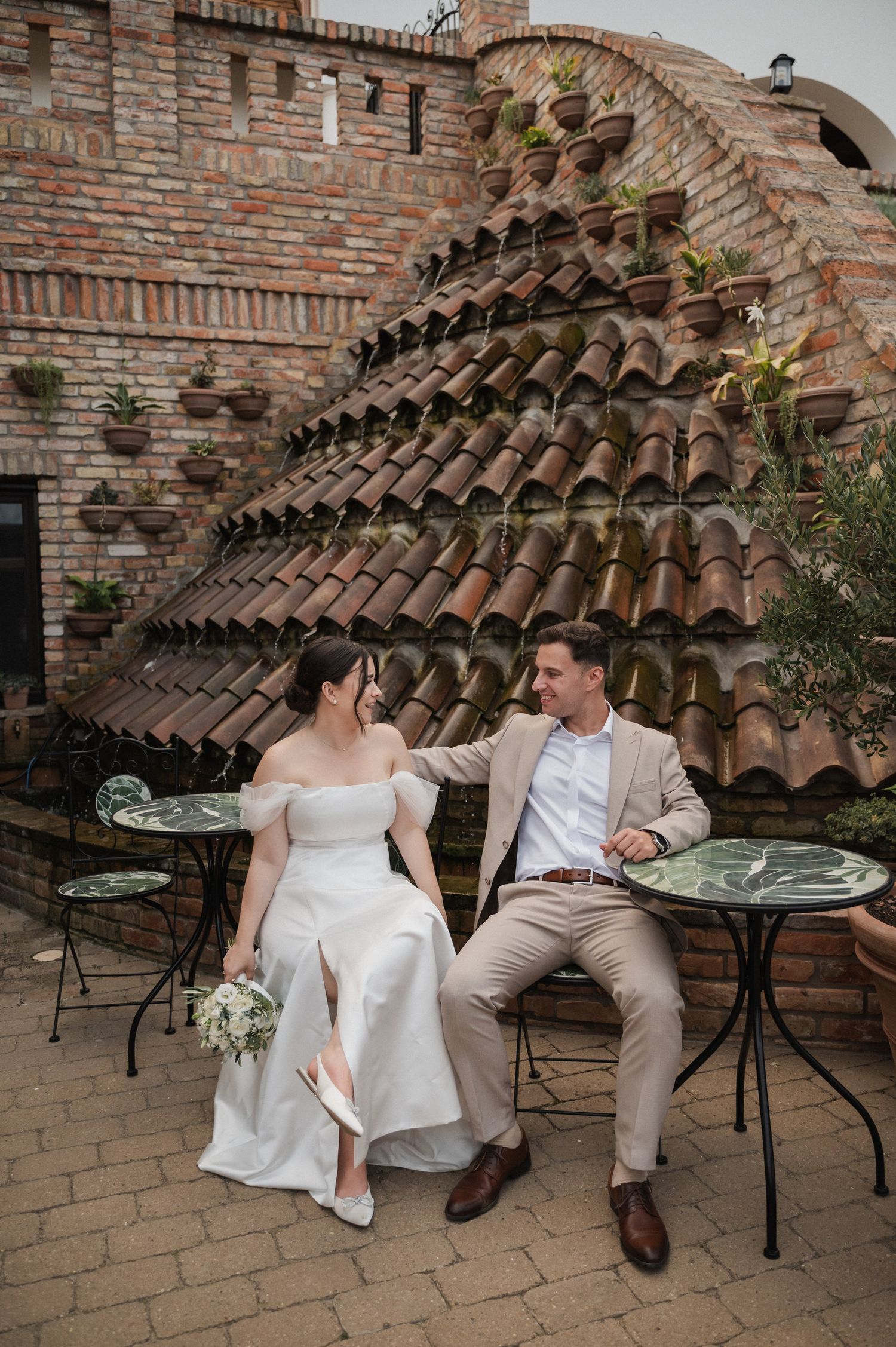A couple in wedding attire sits together on a brick patio with terracotta tile roof details in the background.