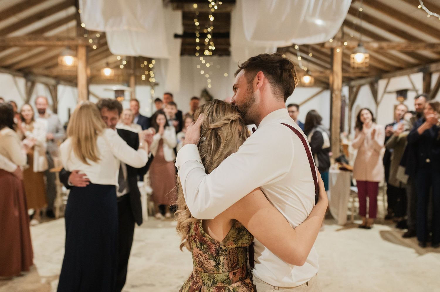 Couples slow dancing at an indoor wedding reception with string lights and wooden beams overhead.