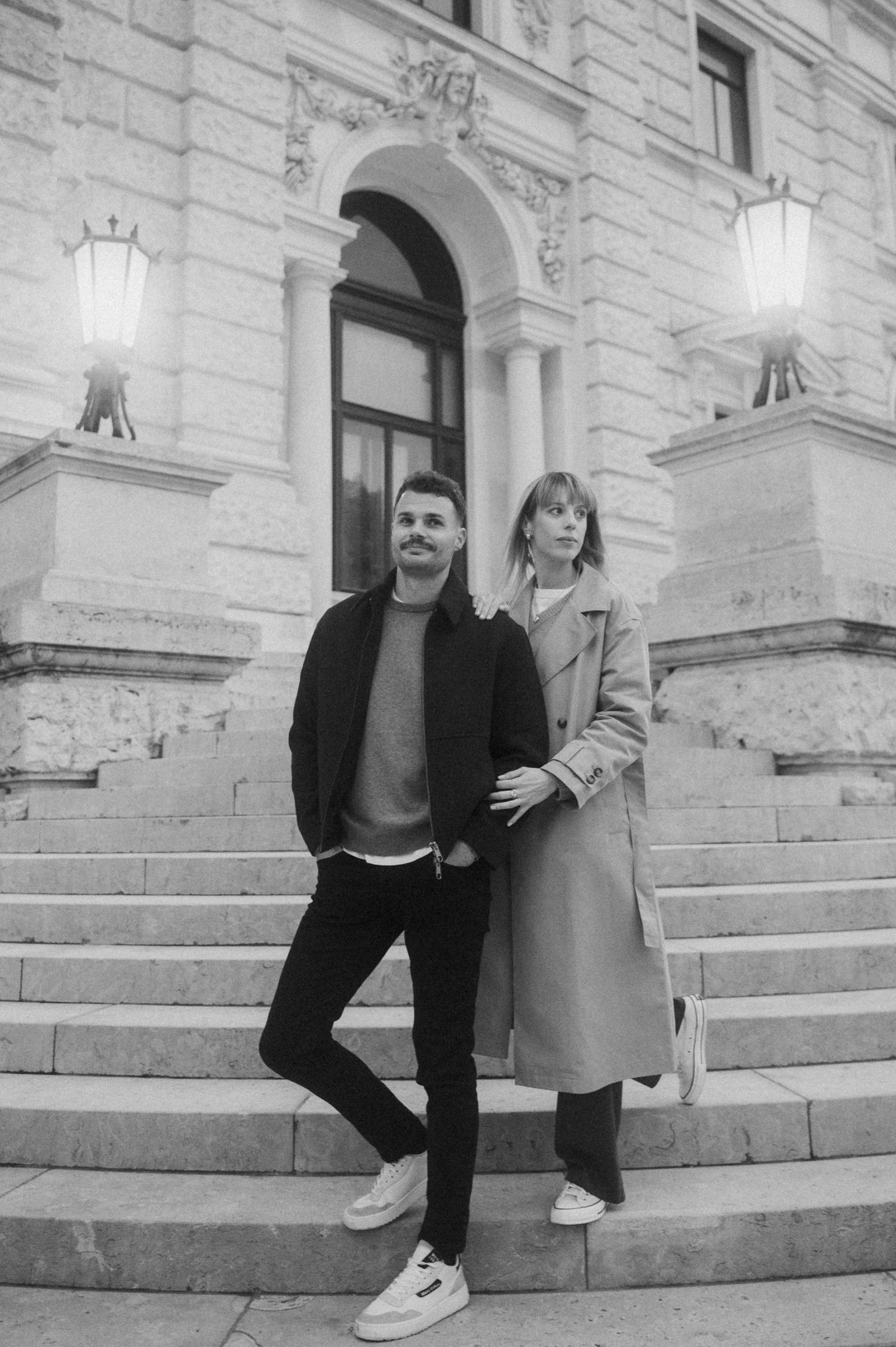 A couple sits together on stone steps outside a historic building with white stone walls and arched windows.