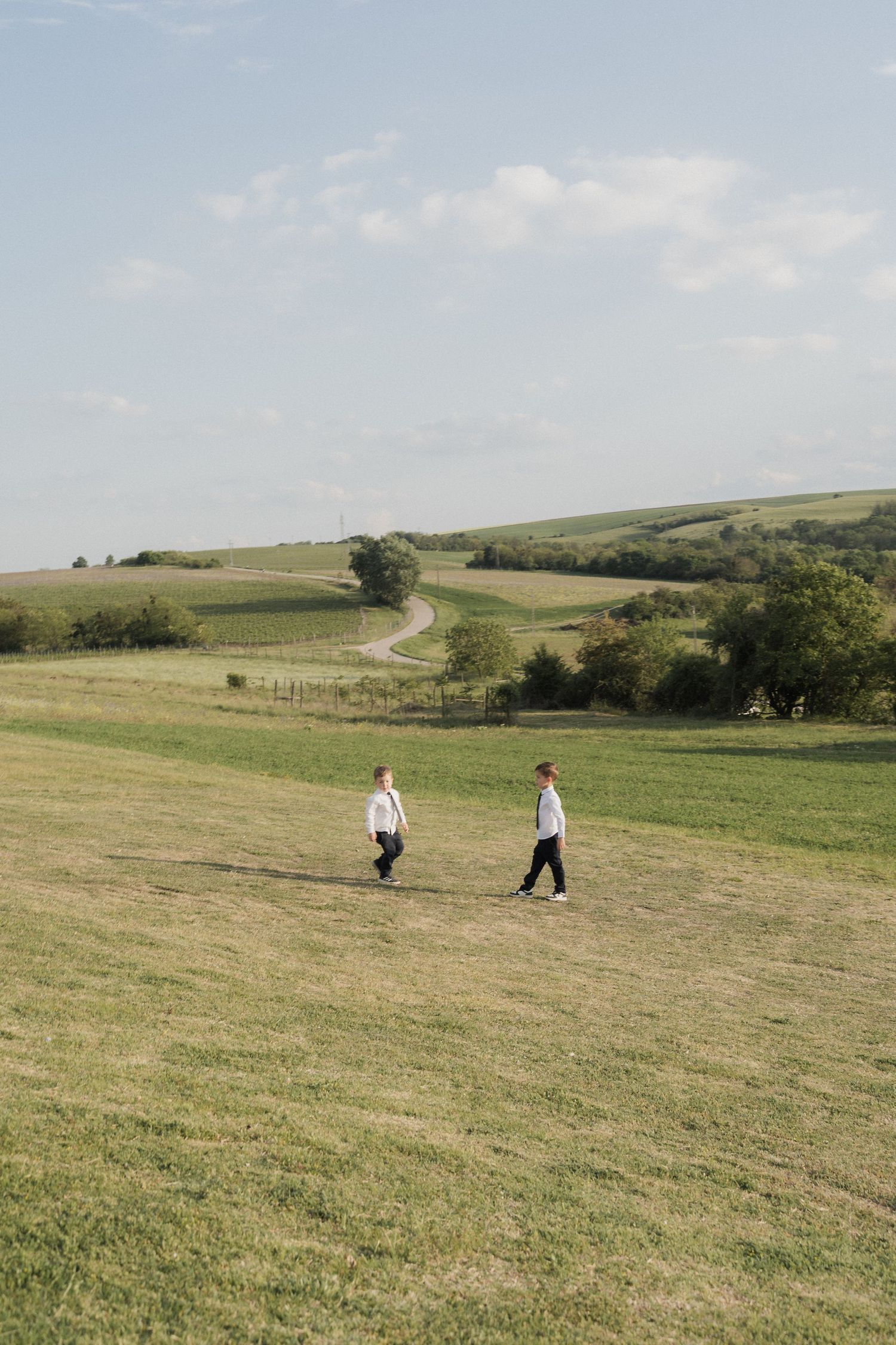 Two men in formal attire walking across a scenic green countryside field under a blue sky.