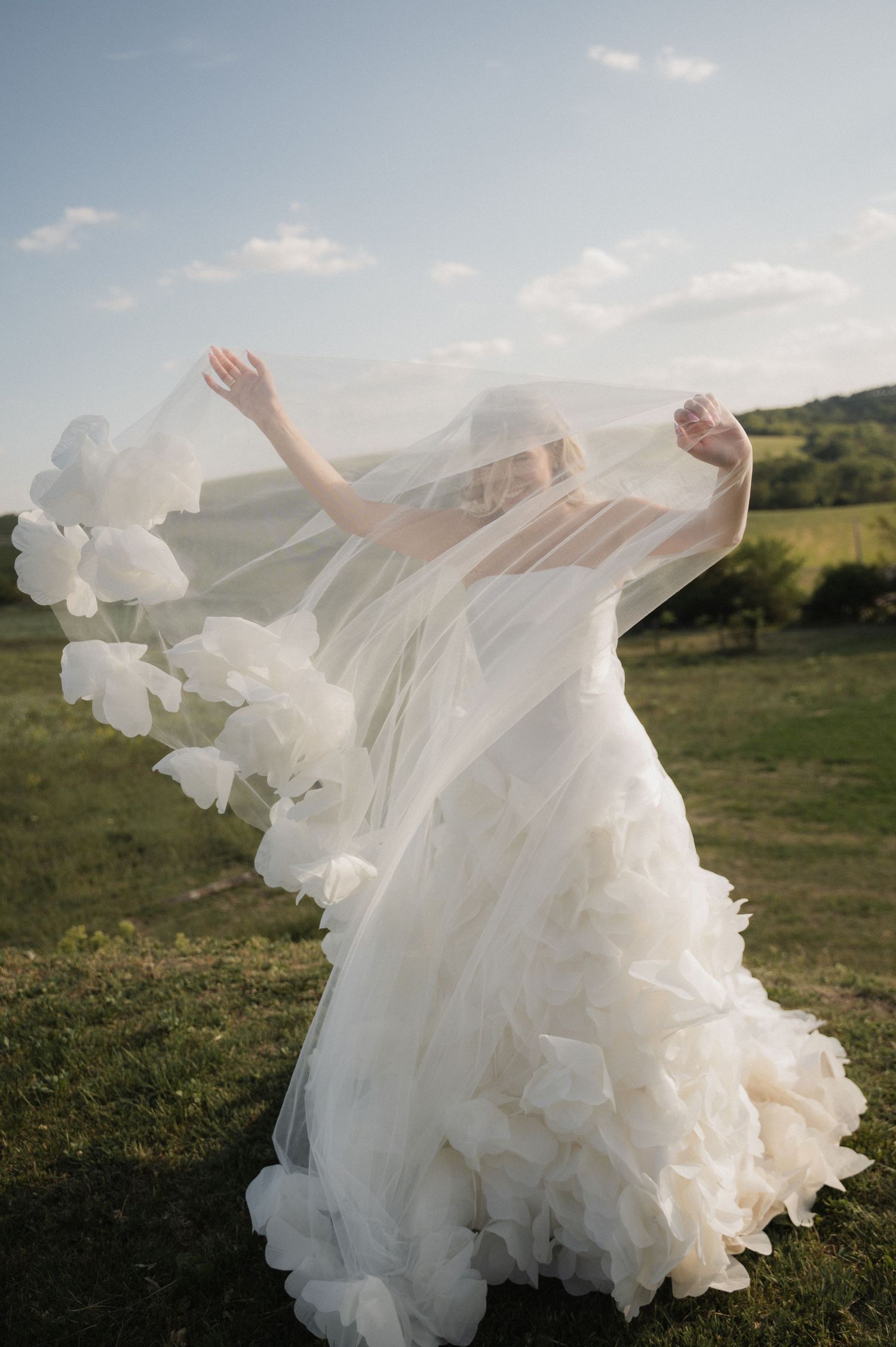 Bride in a ruffled white gown holds her flowing veil wide open in a scenic green countryside under a blue sky.