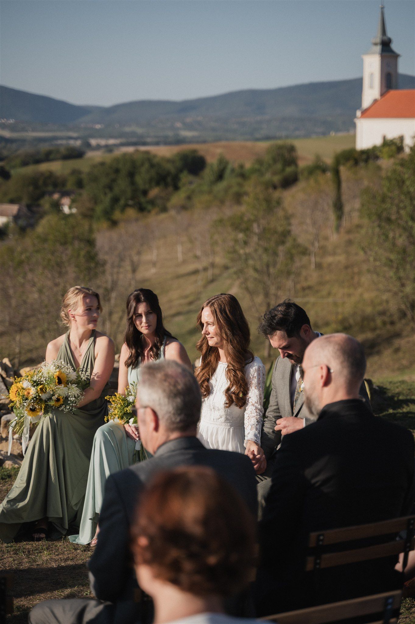Wedding guests seated on wooden benches at an outdoor ceremony with sage green bridesmaid dresses.