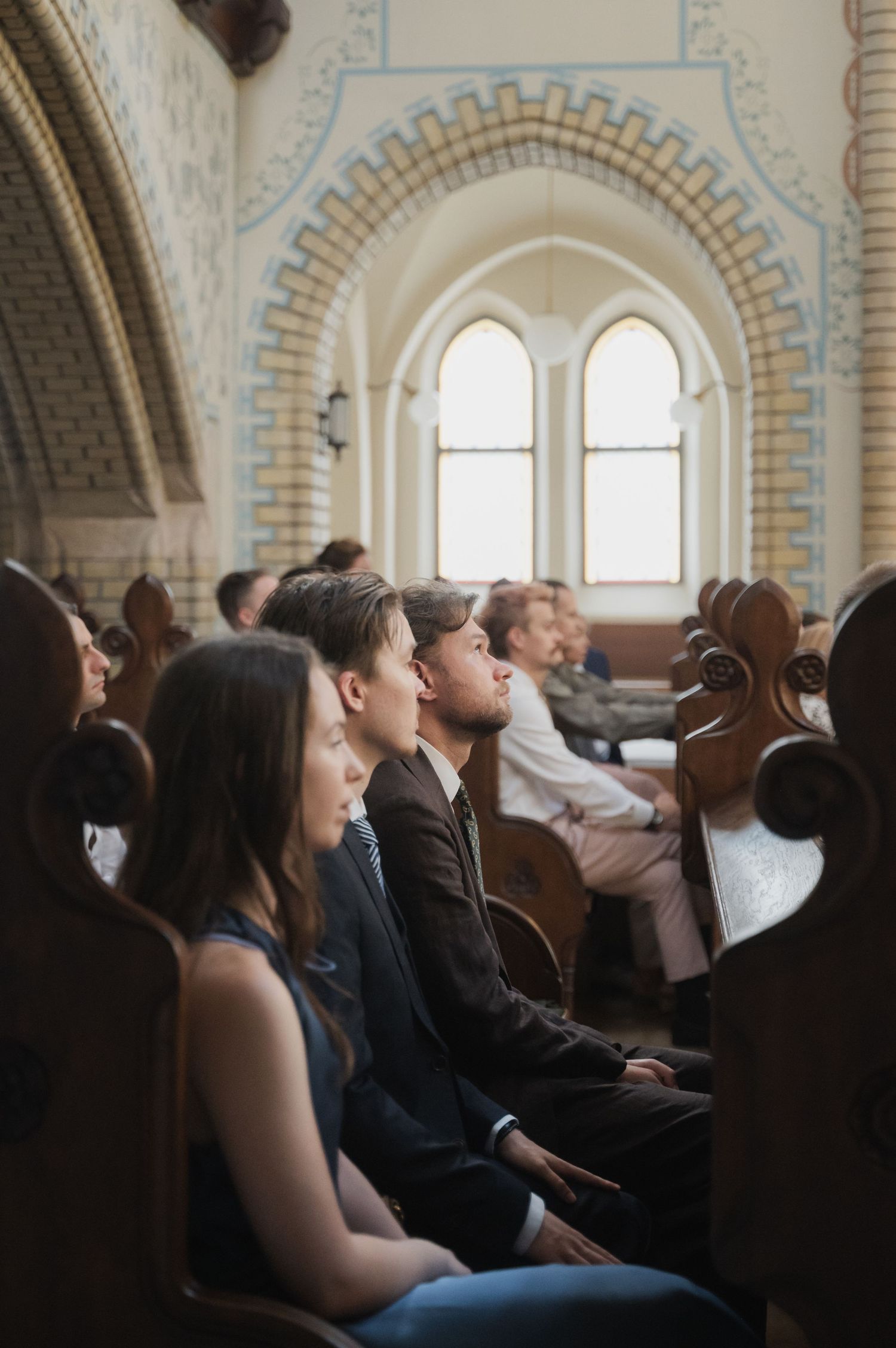 Wedding guests seated in wooden church pews during ceremony inside a beautiful historic church with arched windows.