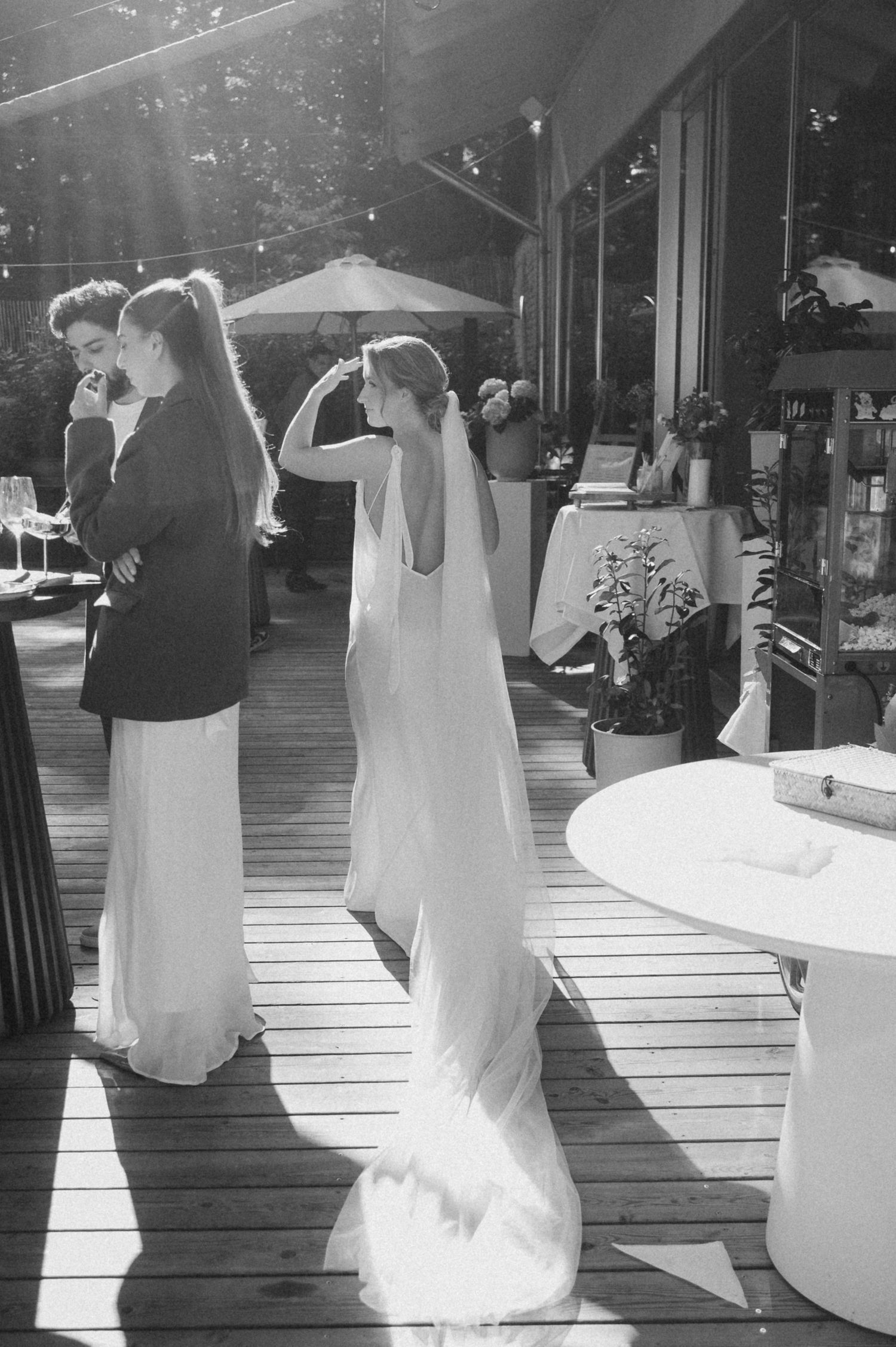 Black and white photo of bride in flowing gown and veil adjusting hair on sunlit outdoor wedding venue deck.