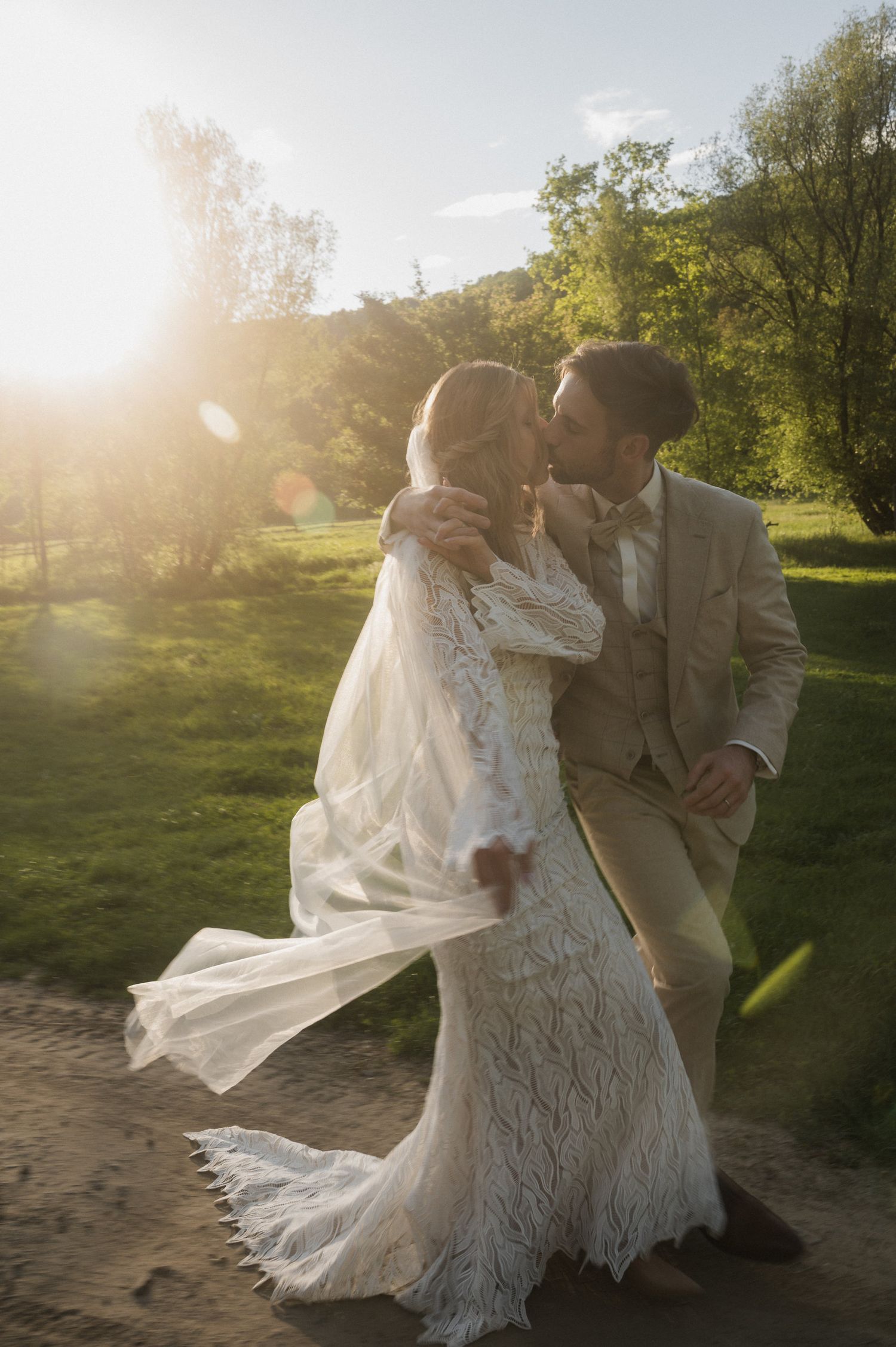 A romantic sunset wedding photoshoot showing a couple dancing in a grassy field with flowing white lace dress and sunflare.