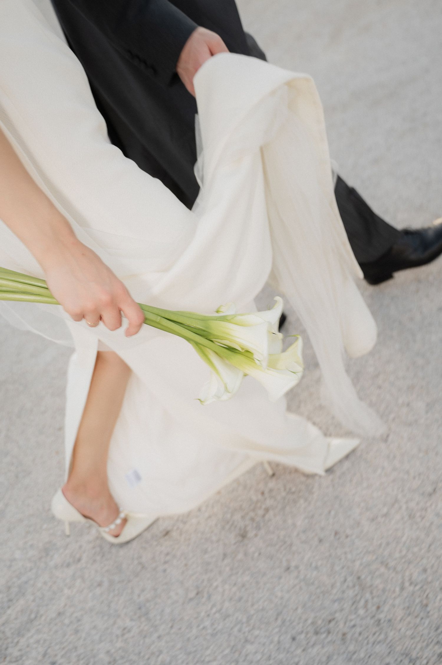 Close-up of wedding dress hem and calla lily bouquet against textured stone ground.