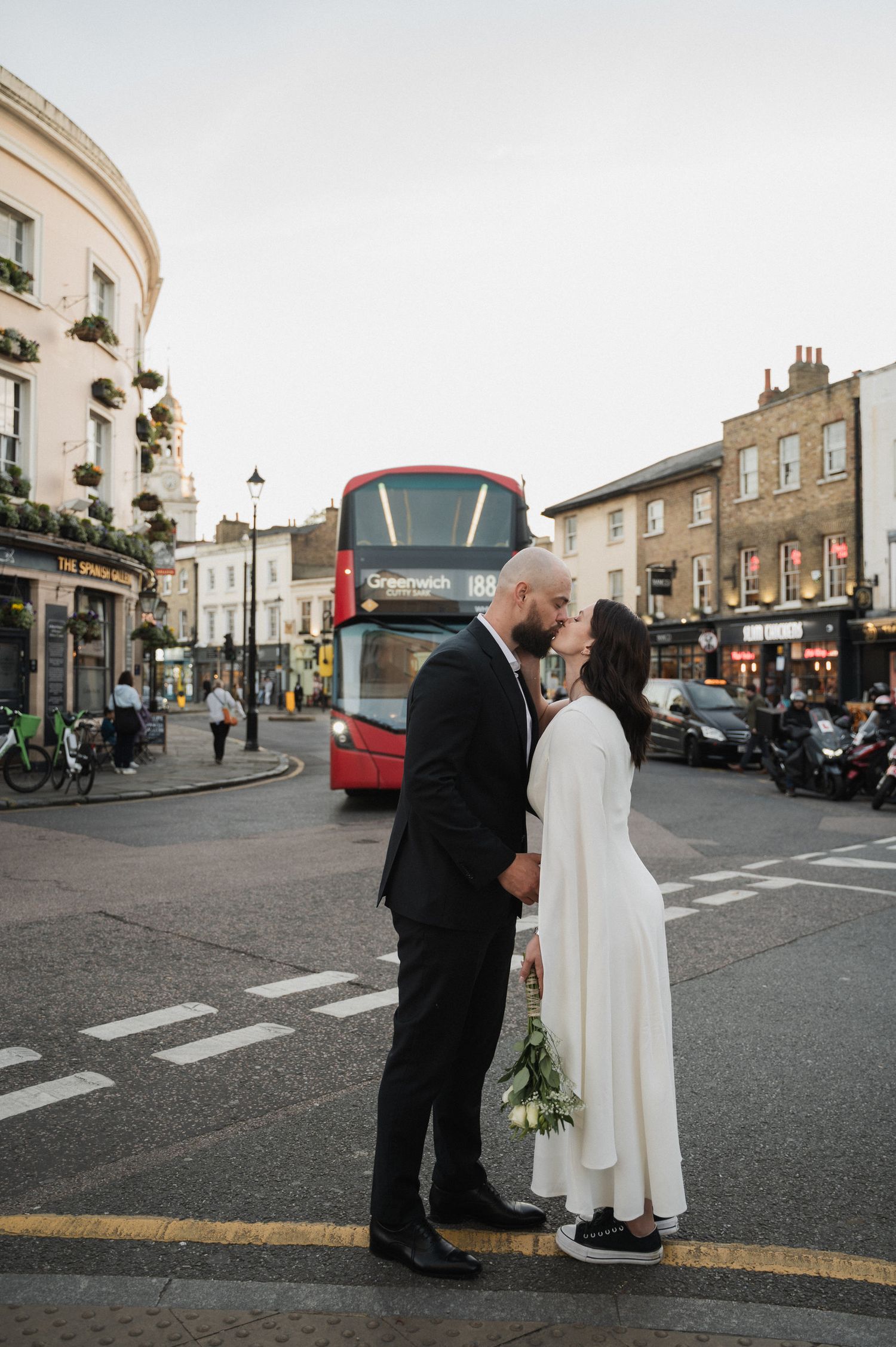 Newlyweds share a moment on a city street with red buses in the background.